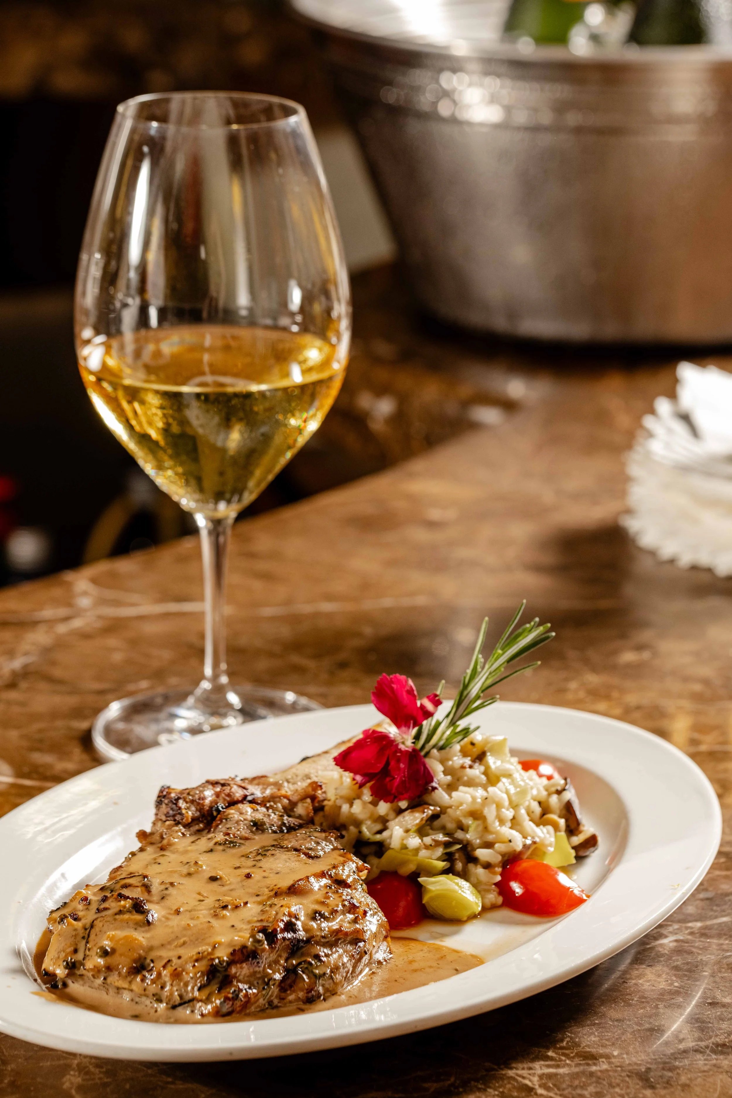A plate with a serving of roasted meat covered in gravy, a side of risotto with vegetables, garnished with a sprig of rosemary and a red edible flower; a glass of white wine in the background.