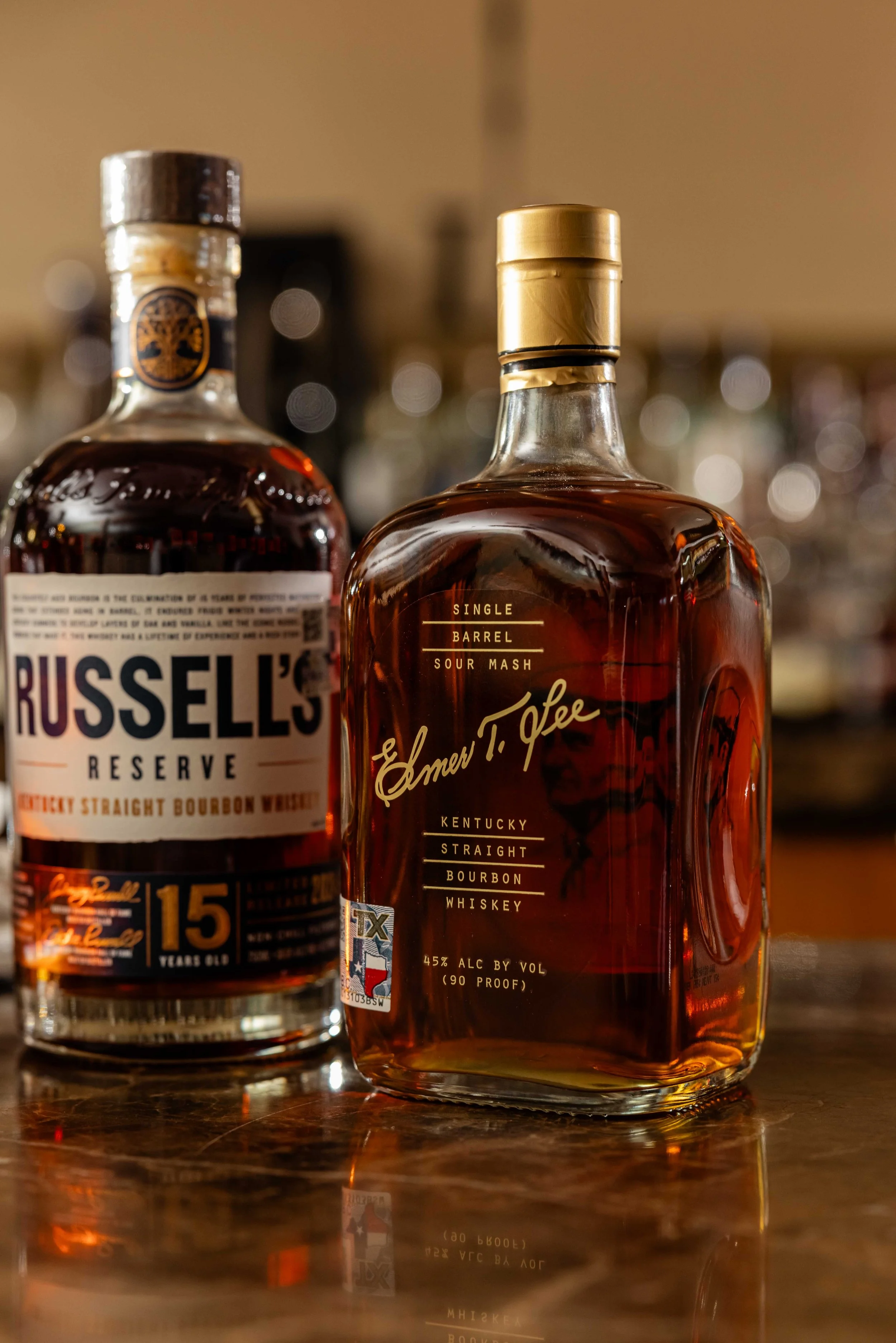 Two bottles of bourbon whiskey on a wooden bar. The bottle in front is labeled Ernesto T. Guee Kentucky Straight Bourbon Whiskey, amber colored liquid, with a gold cap. The bottle behind is Russell's Reserve Kentucky Straight Bourbon Whiskey, 15 year