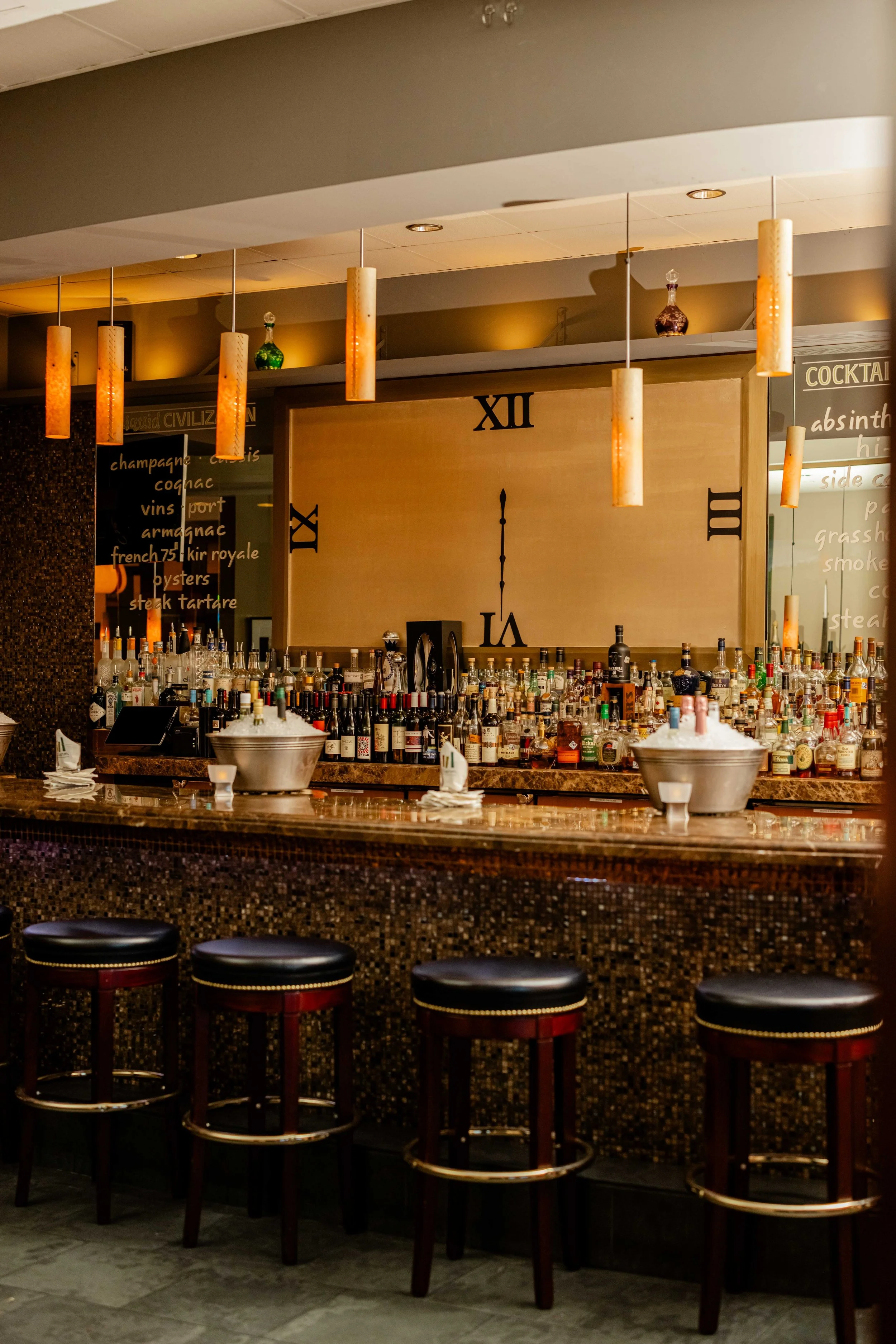 A bar with a large clock on the wall, bottles of alcohol behind the counter, and four bar stools in front of the counter. The lighting is warm and ambient.