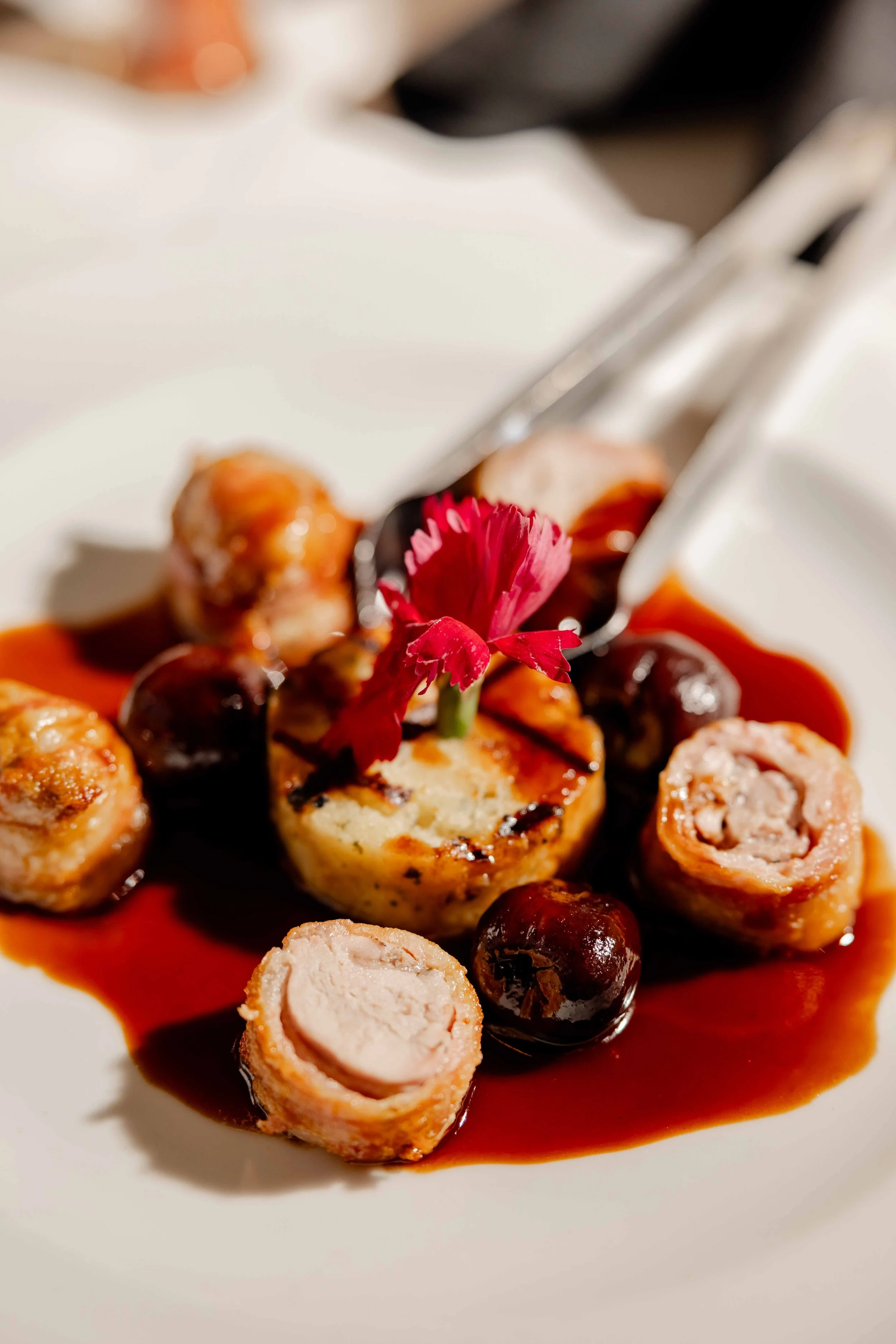 Close-up of stuffed chicken roulades with cherry sauce, garnished with a red edible flower, on a white plate.