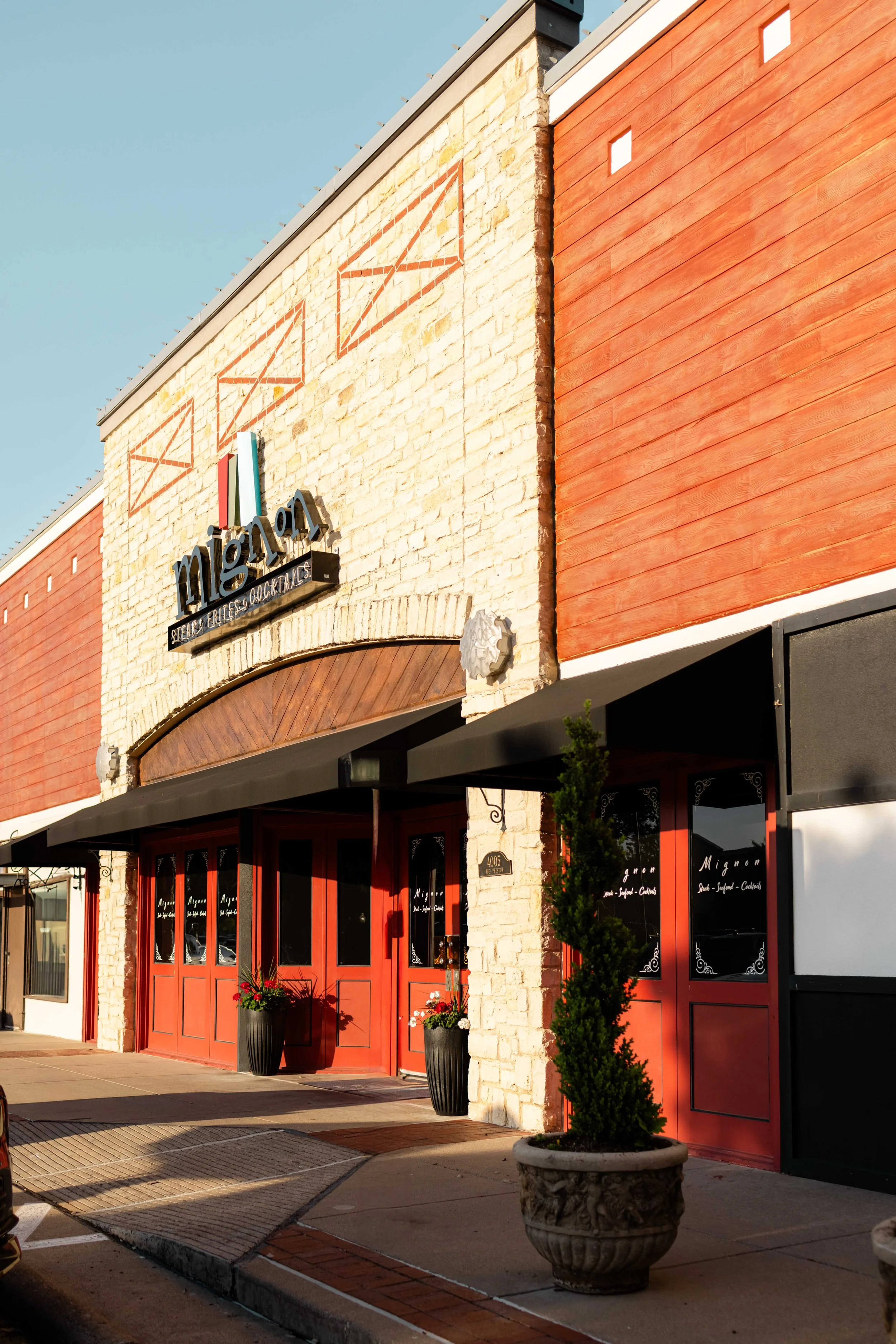 Exterior view of a fine dining restaurant named Mignon with a sign, red doors, potted plants, and a mix of brick and wood siding on the building.