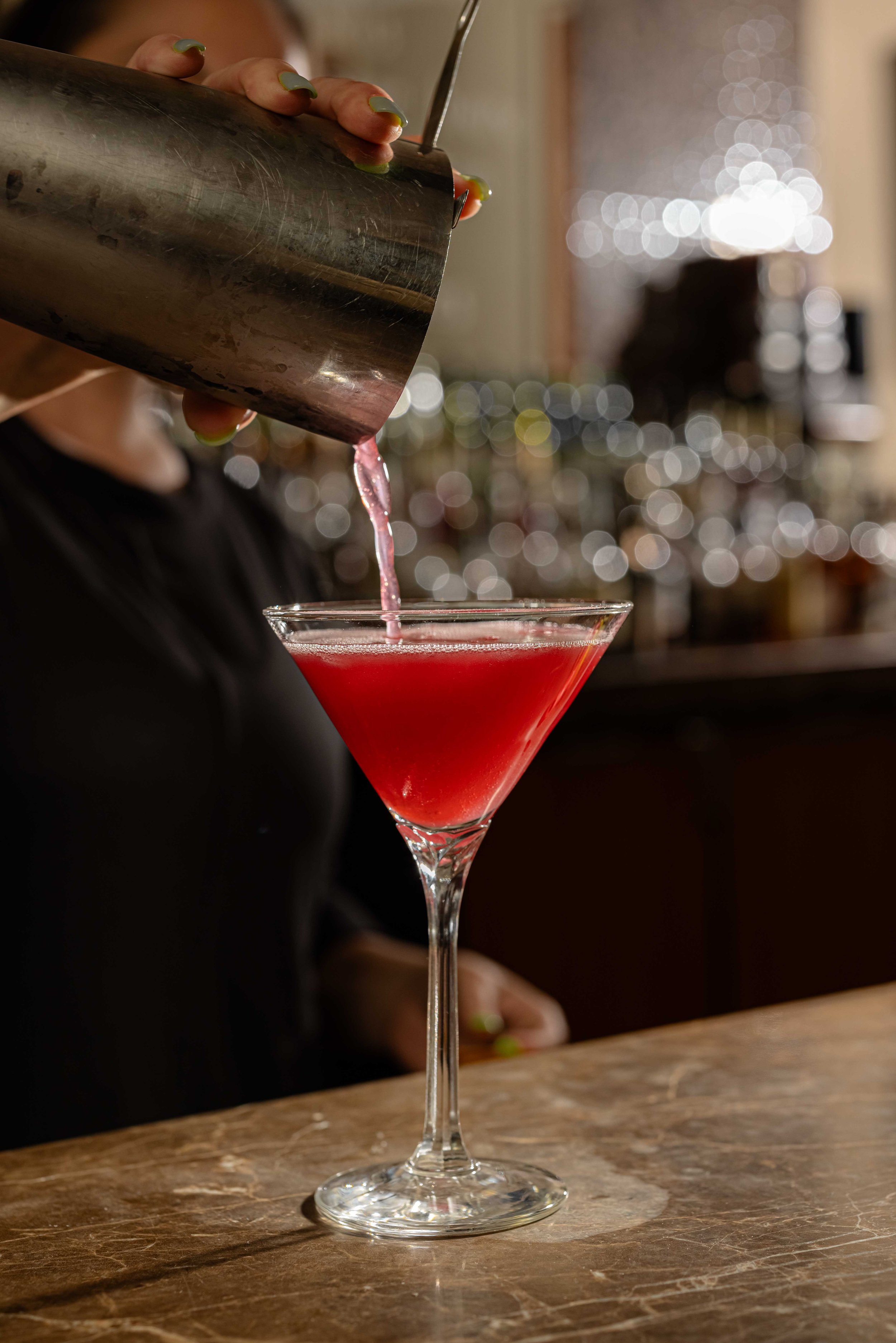 A person pouring a pink cocktail into a martini glass at a bar, with a blurred background of bar lights.