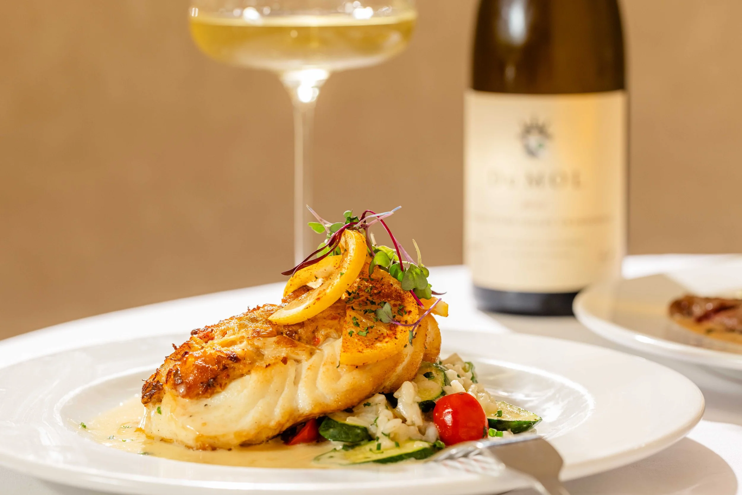 A plated serving of pan roasted halibut fish topped with lemon slices and microgreens, accompanied by a vegetable risotto, with a glass of white wine and a bottle in the background.