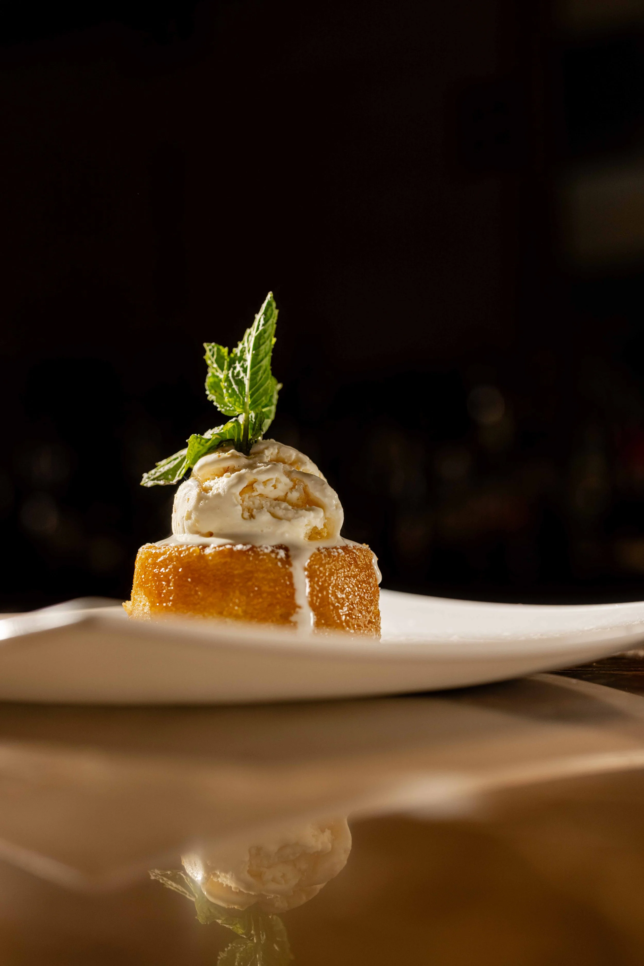 A piece of dessert topped with a scoop of ice cream, garnished with a sprig of mint, on a white plate with a dark background.