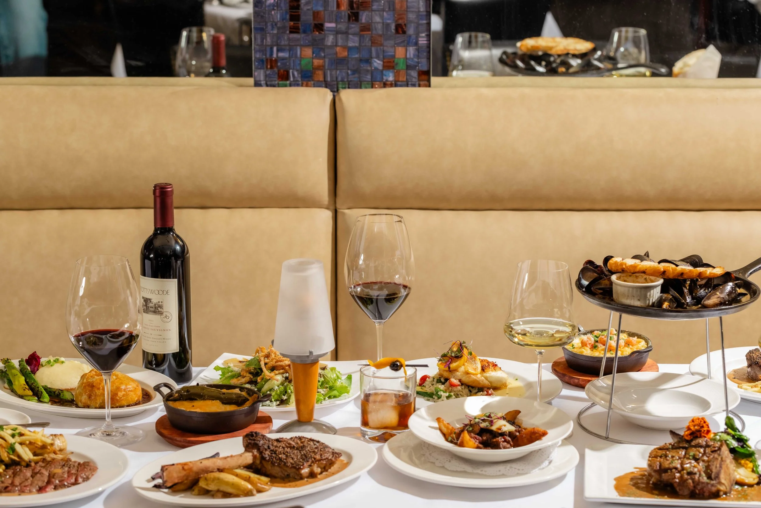 Restaurant table with various wines and dishes, including seafood, steak, salads, and desserts, arranged for a meal.