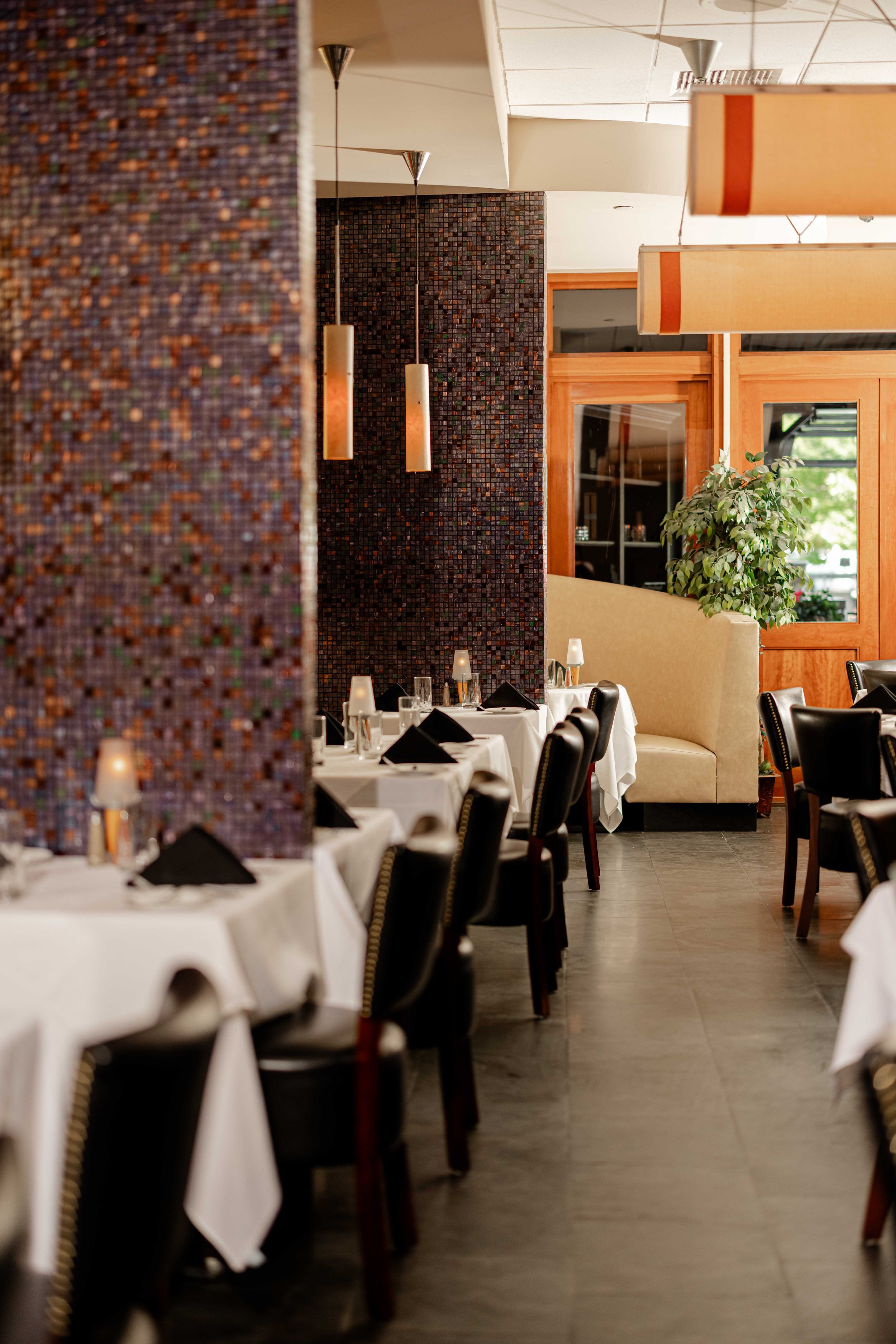 Empty restaurant dining room with tables set for service, black chairs, white tablecloths, black napkins, and small candles; interior decor includes textured walls, hanging pendant lights, and a large potted plant near windows.