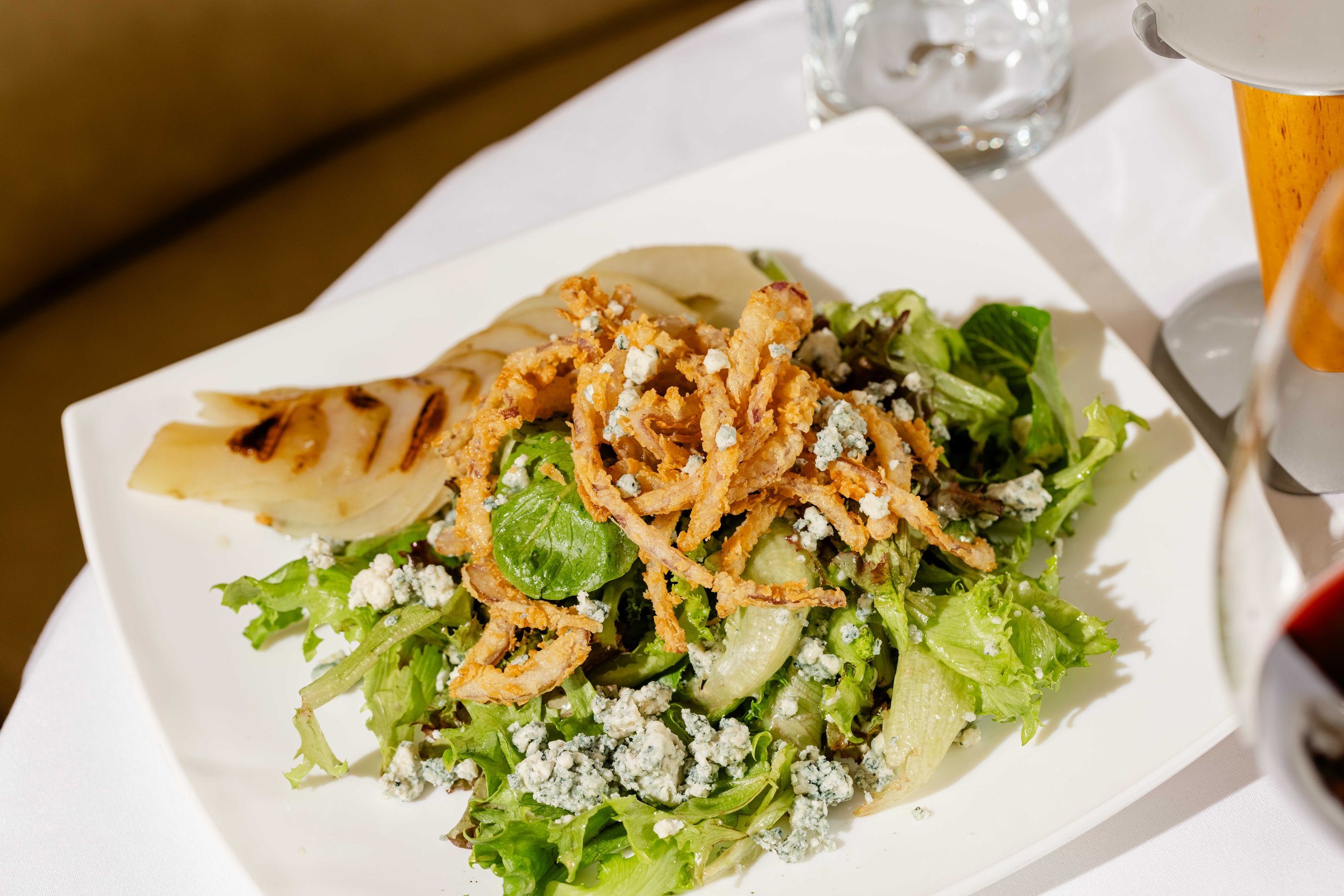 Fresh green salad with sliced grilled pear, blue cheese crumbles, and crispy fried onions on a white plate.