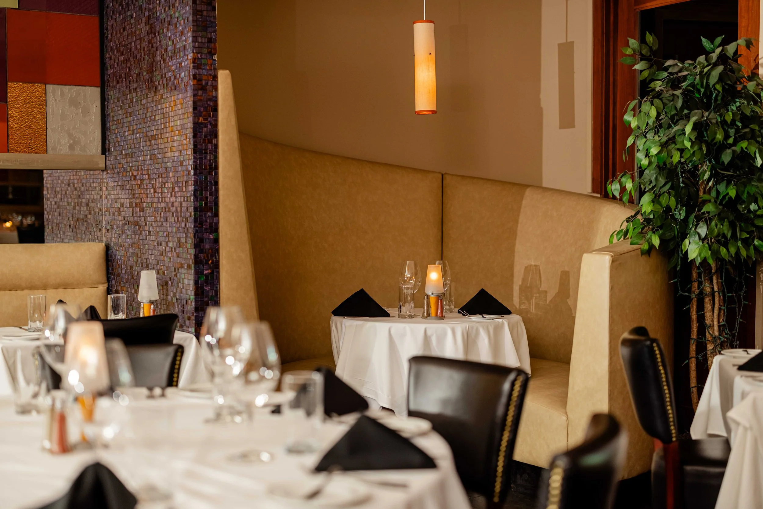 Elegant restaurant dining area with white tablecloths, black napkins, glasses, candles, and booth seating, decorated with warm lighting and a potted plant.