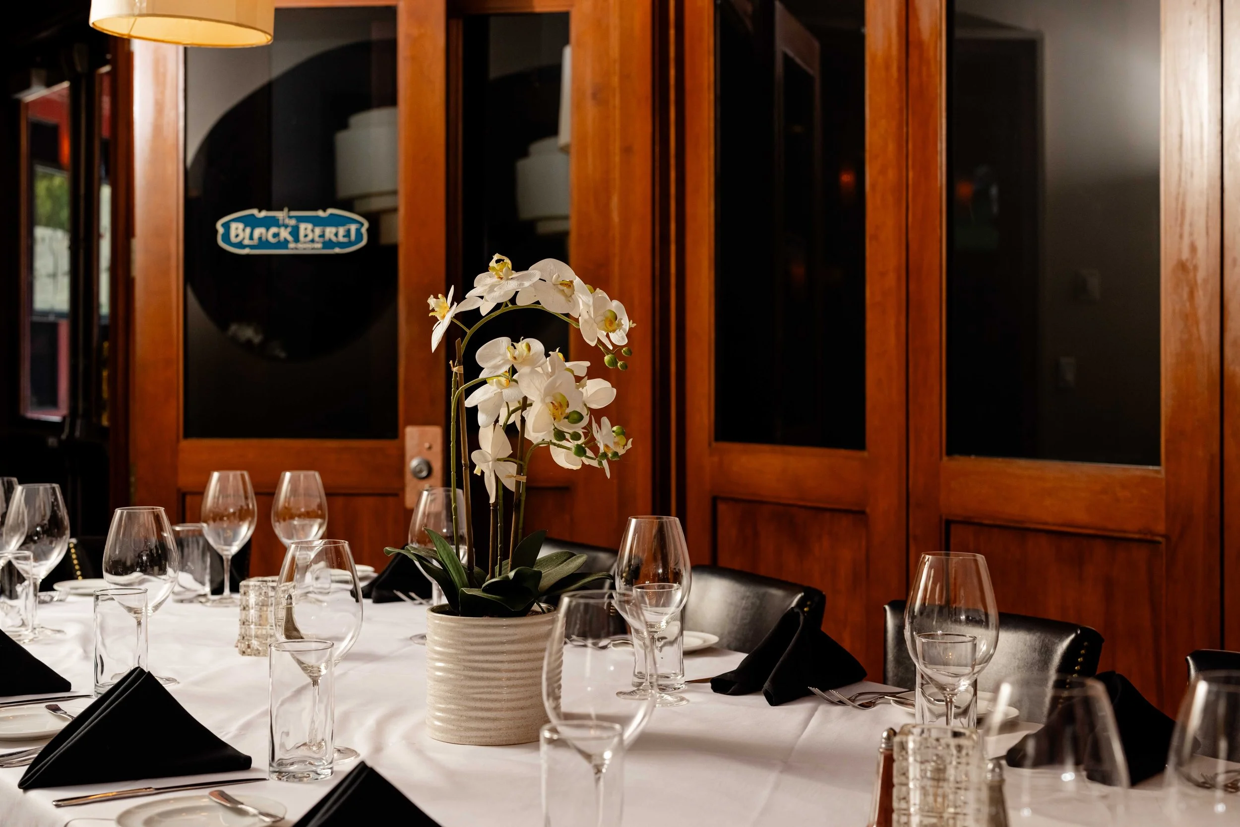 Elegant dining table set with wine glasses, black napkins, and a white flower centerpiece in a restaurant with wooden paneled walls.