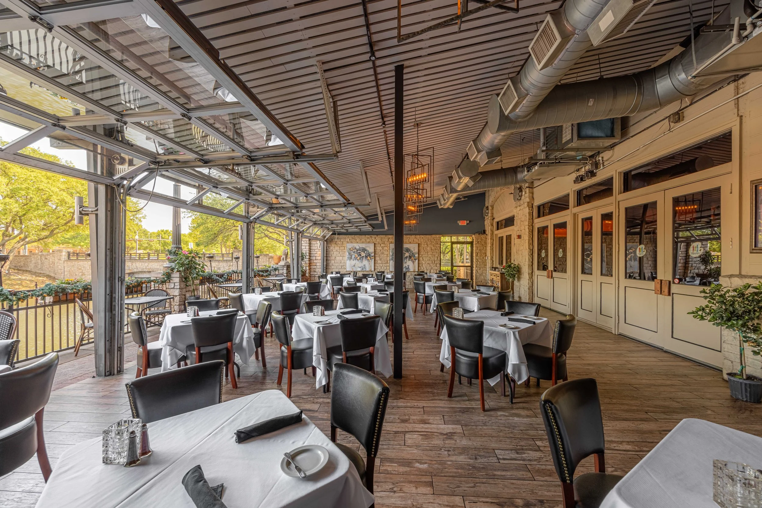 Empty restaurant dining area with white tablecloth-covered tables, black chairs, and open garage-style windows, decorated for a meal.