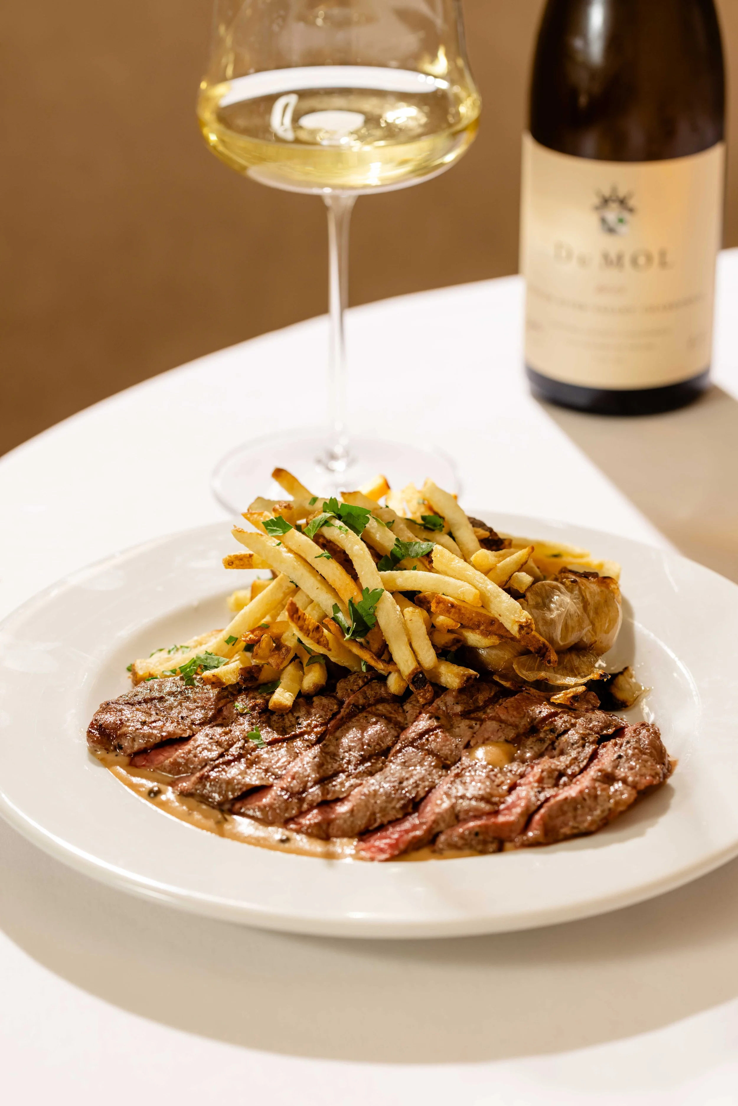 A plate of sliced steak with fries and herbs, a glass of white wine, and a bottle of wine on a table.