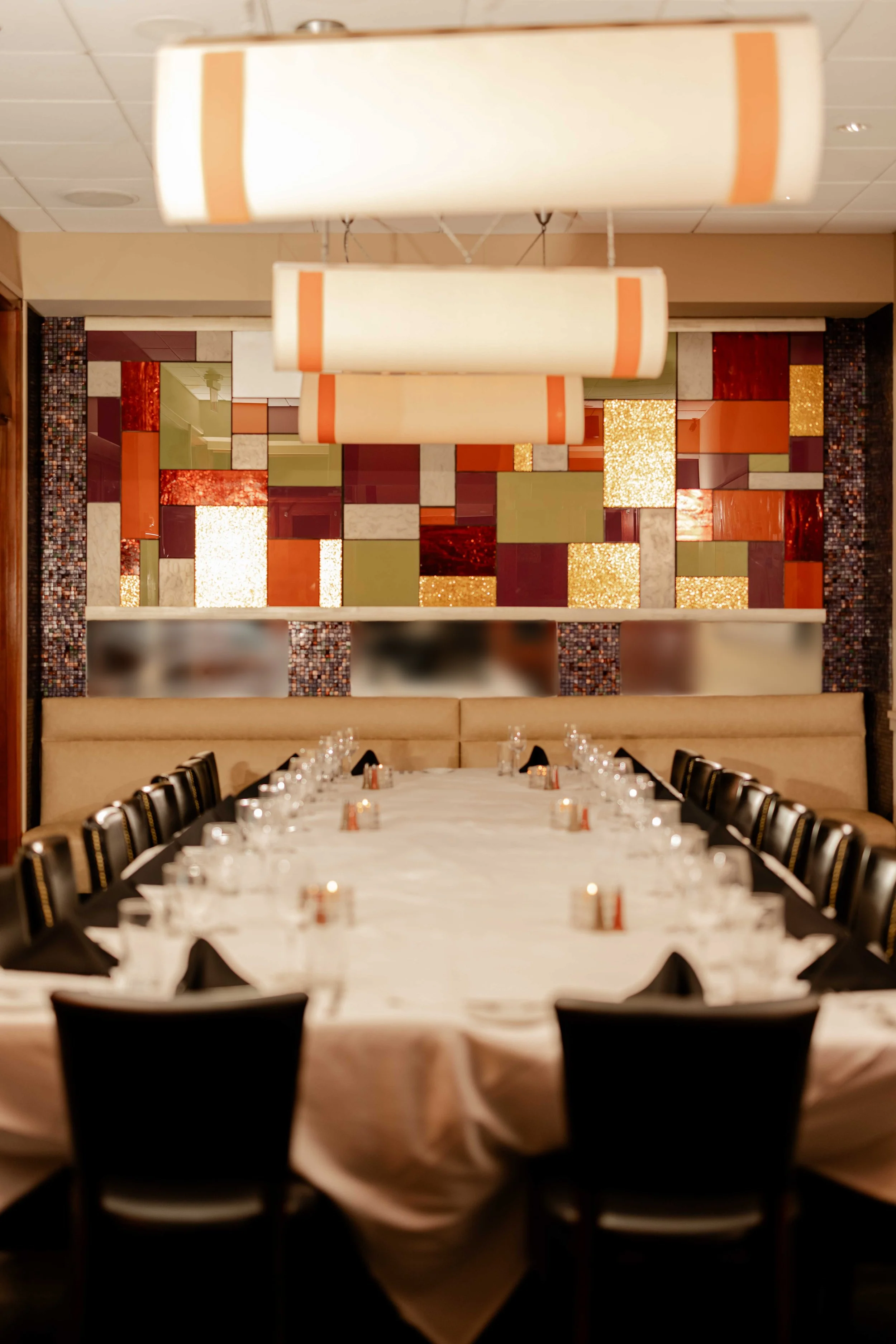 A banquet room with a long, oval dining table covered with a white tablecloth, set with glasses, napkins, and small candles. The room features a colorful, abstract mosaic wall and large hanging light fixtures.
