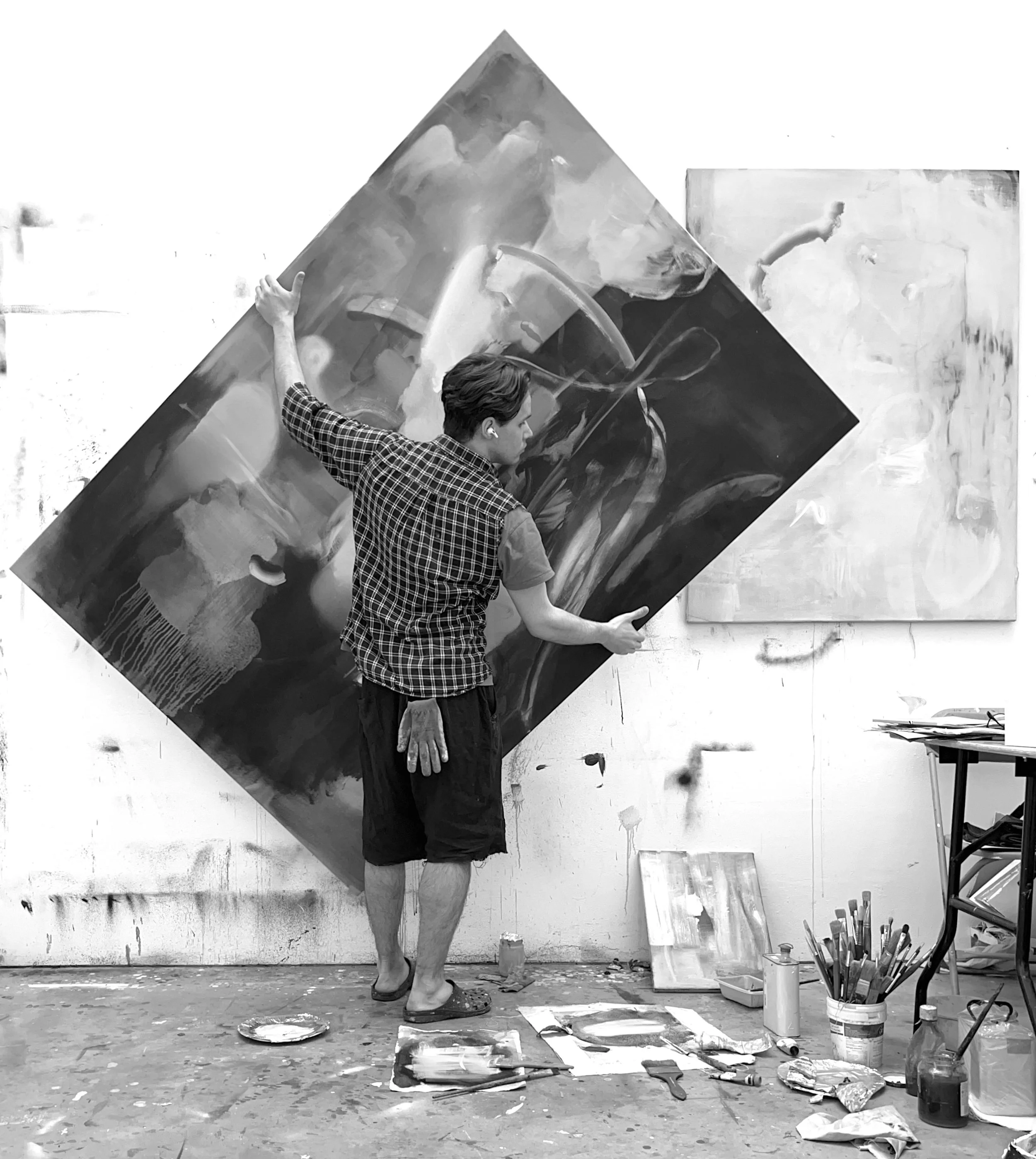 Jan Valik, an artist in his studio setting working on a large abstract painting. The artist is wearing a checkered shirt and shorts, holding a paintbrush, with various paints and art supplies scattered around.
