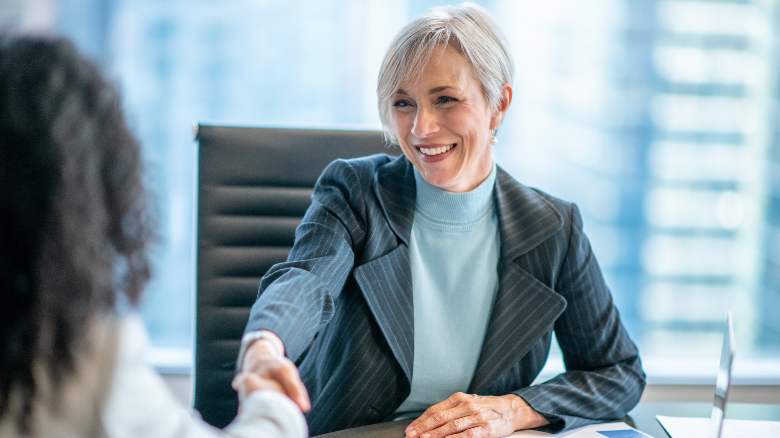 A professional woman with short gray hair smiling and shaking hands with a person in an office setting.
