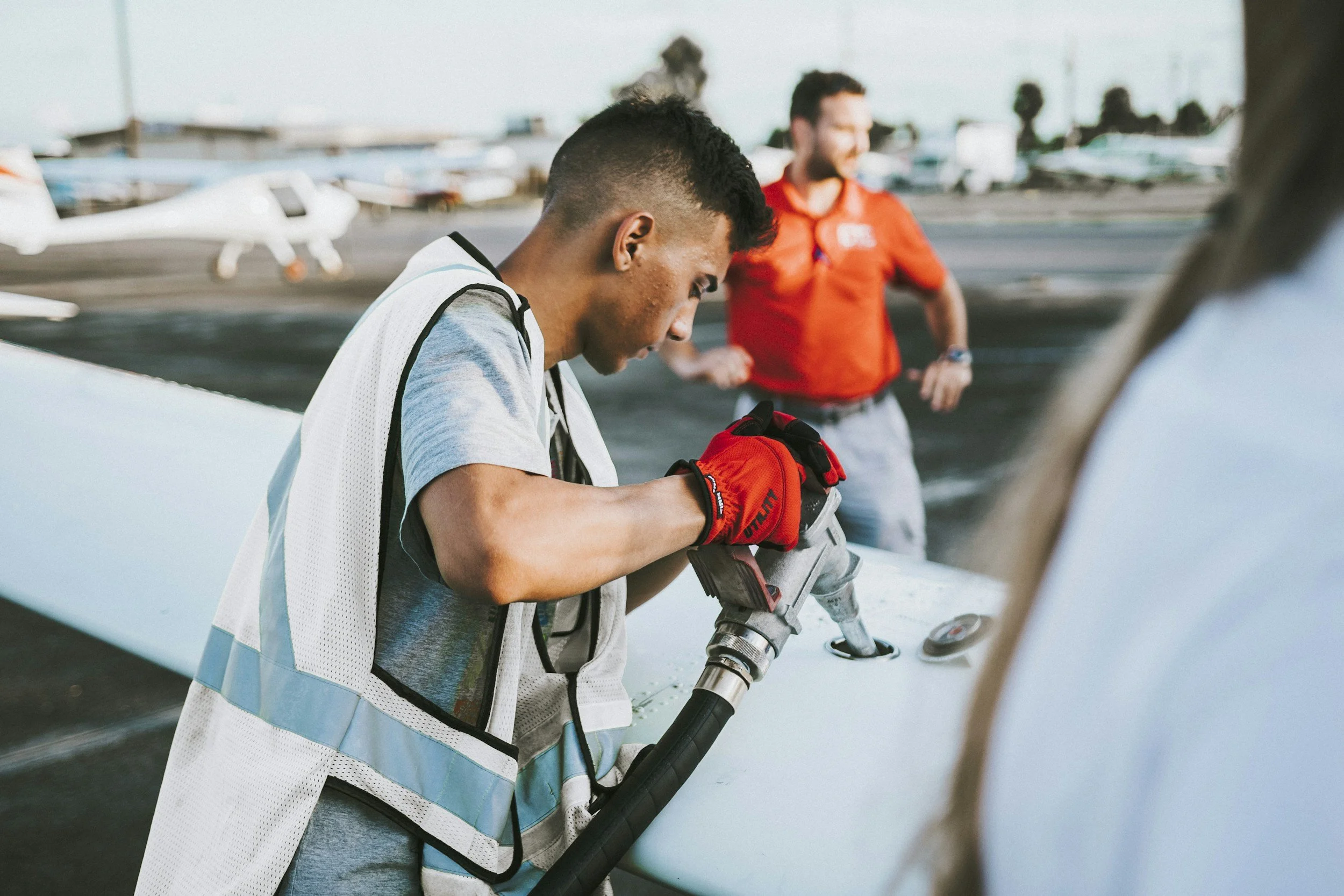 A man wearing a safety vest and gloves working on a small aircraft with a power tool, while another man in a red shirt stands in the background at an airfield with several small planes parked.