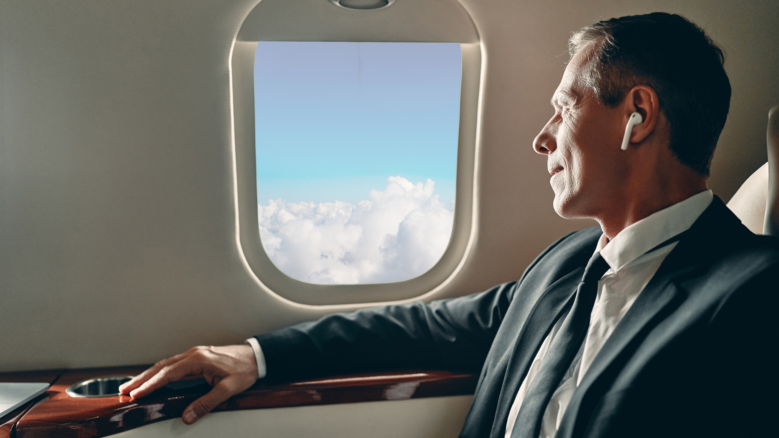 A man wearing a suit and tie, sitting on an airplane seat, looking out the window at clouds and blue sky with a slight smile, while wearing wireless earbuds.