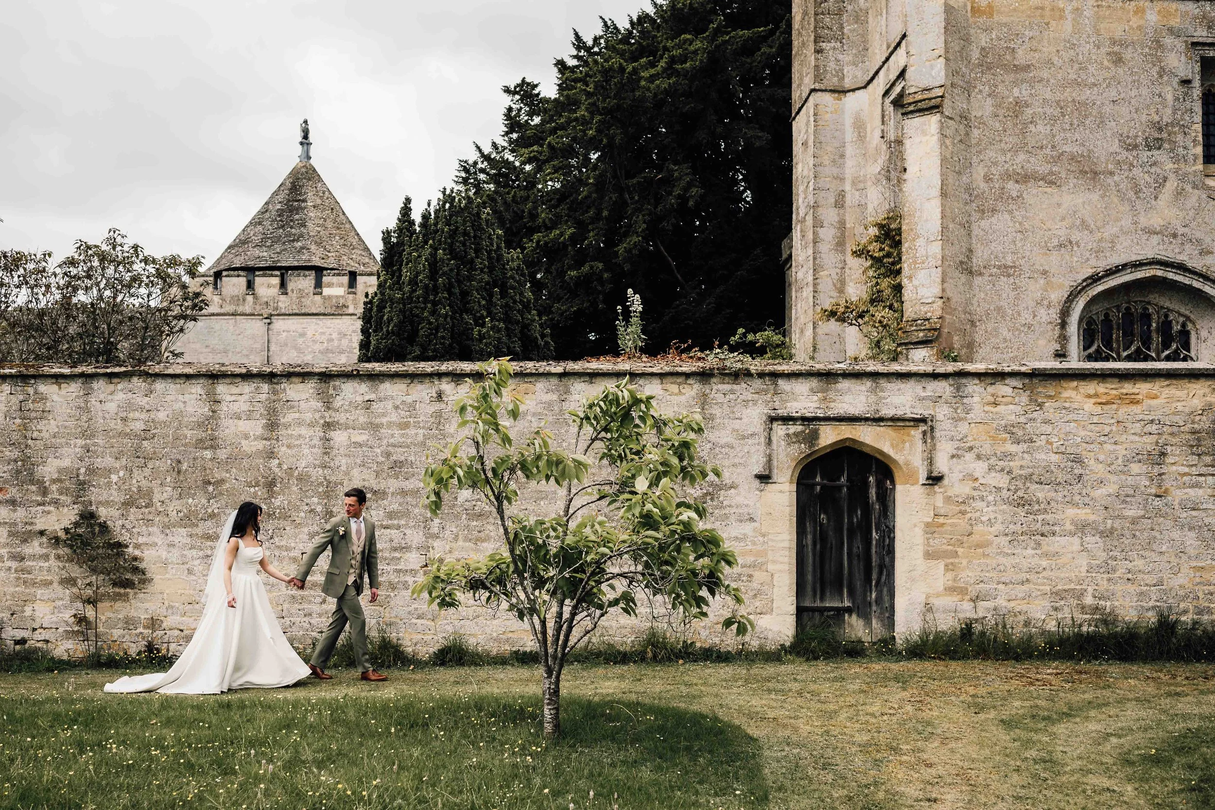 A bride and groom holding hands in front of an old stone building with a tower and a small arched window, a small tree in the foreground, cloudy sky overhead.
