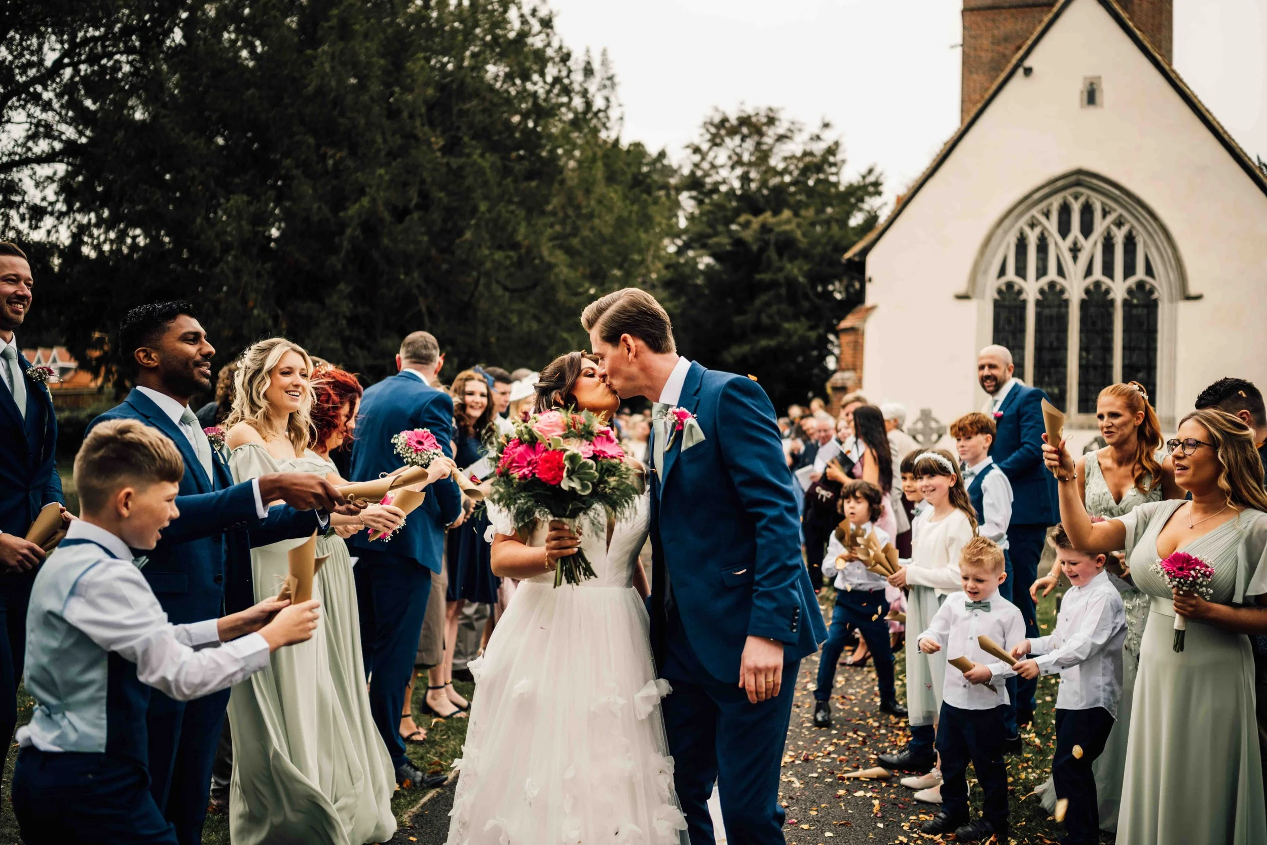 A newlywed couple sharing a kiss surrounded by wedding guests outside a church, with friends and family holding flowers and programs.