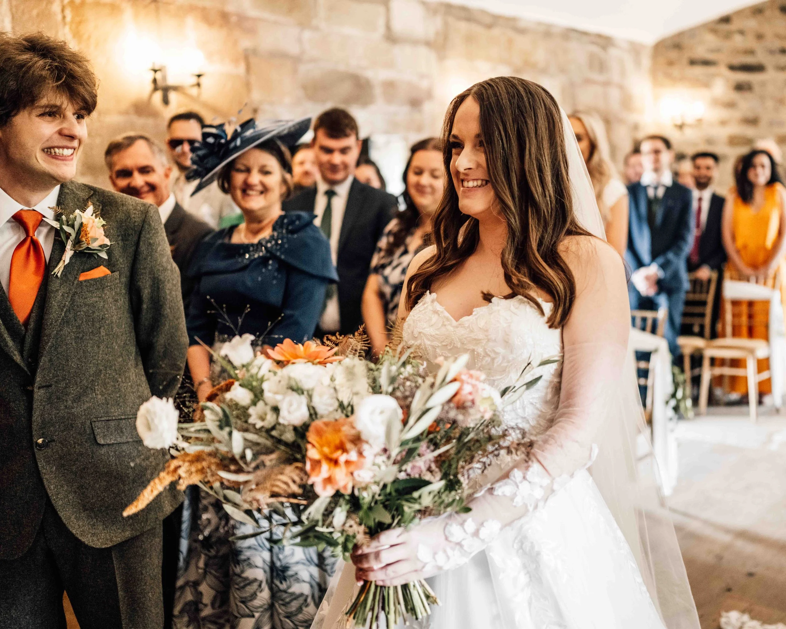 Bride in a white wedding dress holding a large bouquet of flowers, smiling at the groom during their wedding ceremony. Guests in formal attire are gathered in the background inside a stone-walled room.