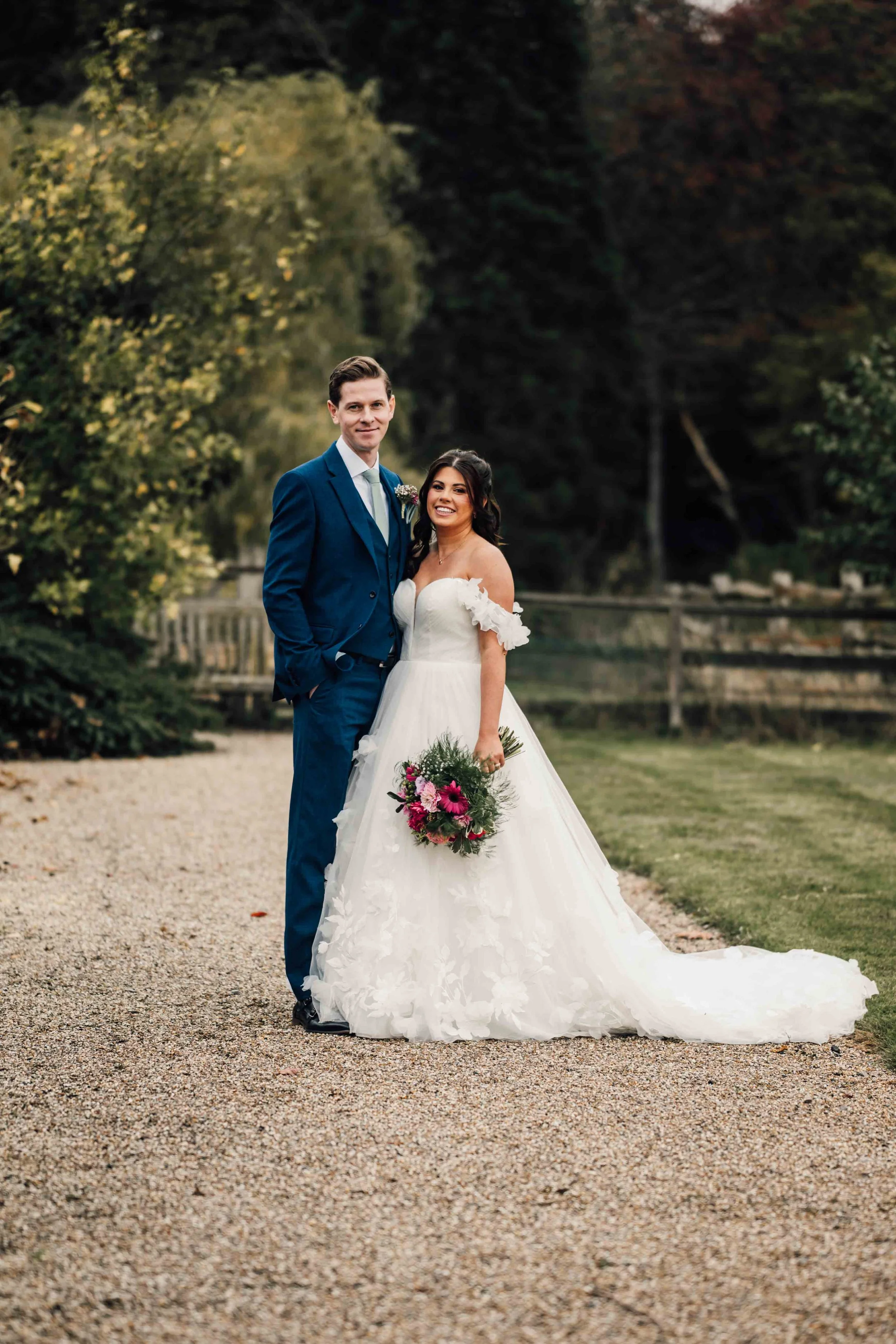 A bride and groom standing together outdoors on a gravel path surrounded by trees, with the bride holding a bouquet of pink and purple flowers.