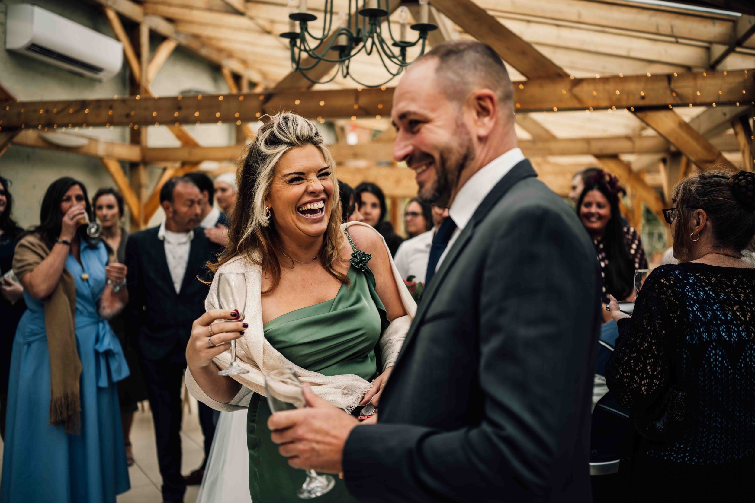 A group of people at a wedding reception inside a rustic wooden venue, with a woman in a green dress laughing and holding a drink, and a man in a suit smiling nearby, surrounded by other guests.