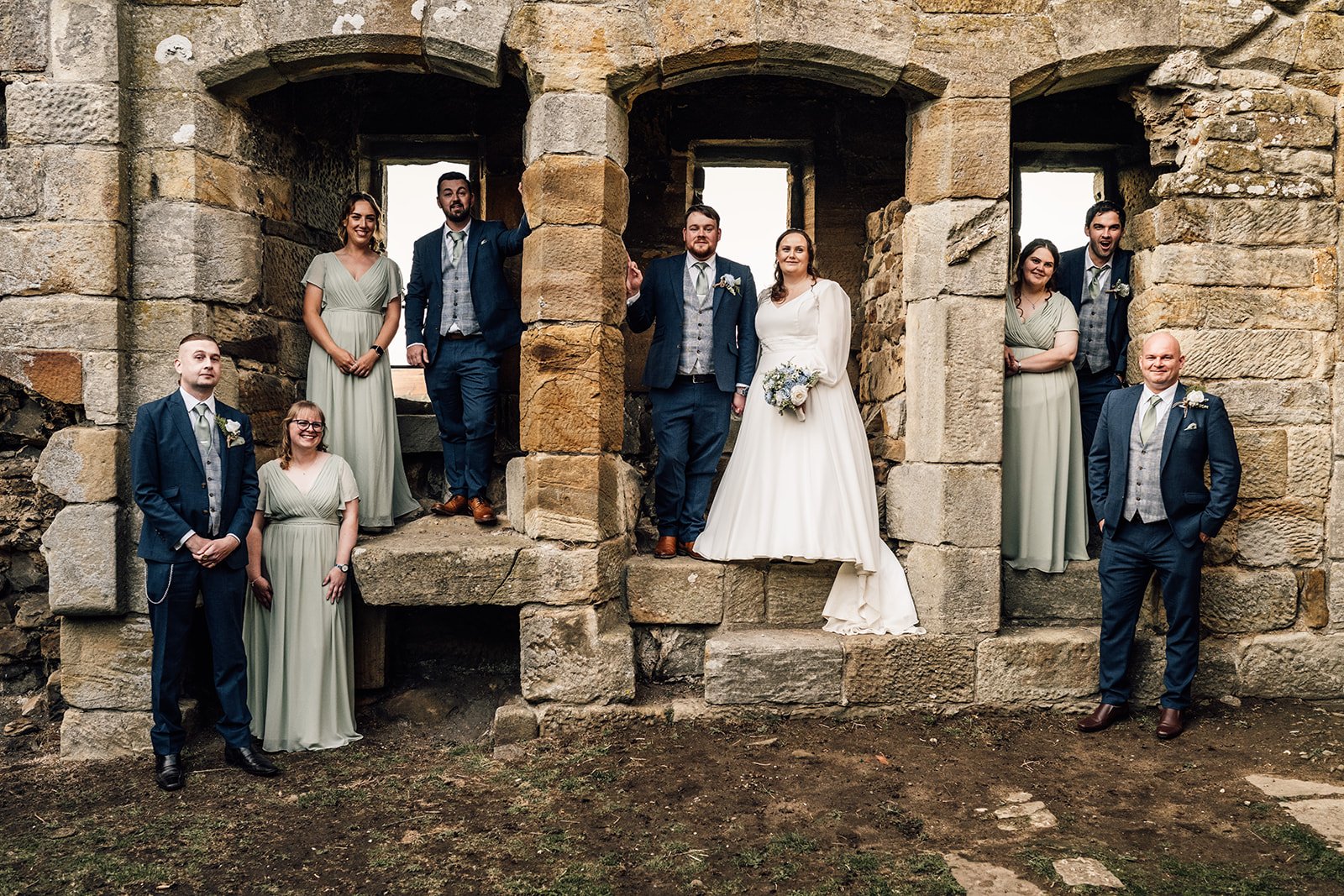 Group of eight people at a wedding photo shoot in front of a stone ruin. The bride is in a white gown holding a bouquet, and the groom and others are in suits and dresses.