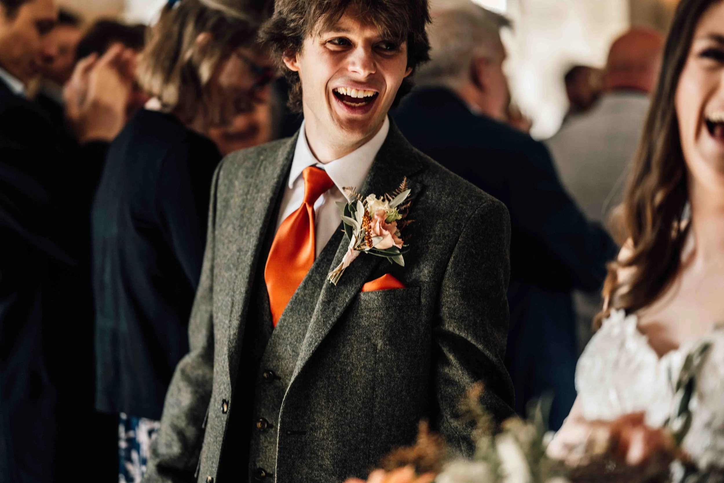Young man in a gray suit with an orange tie and boutonnière, smiling and laughing at a wedding reception with other guests in the background.