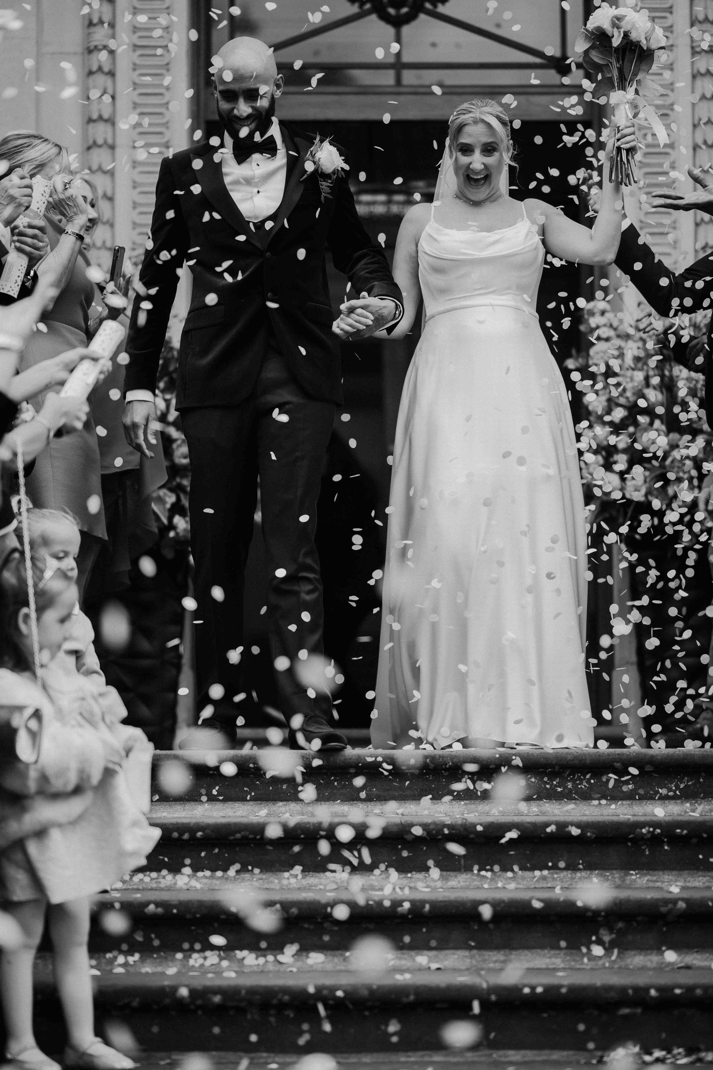 A black and white photo of a newlywed couple walking down stairs after their wedding ceremony, surrounded by guests throwing confetti. The bride is holding a bouquet and has a joyful expression, while the groom is smiling and holding her hand.