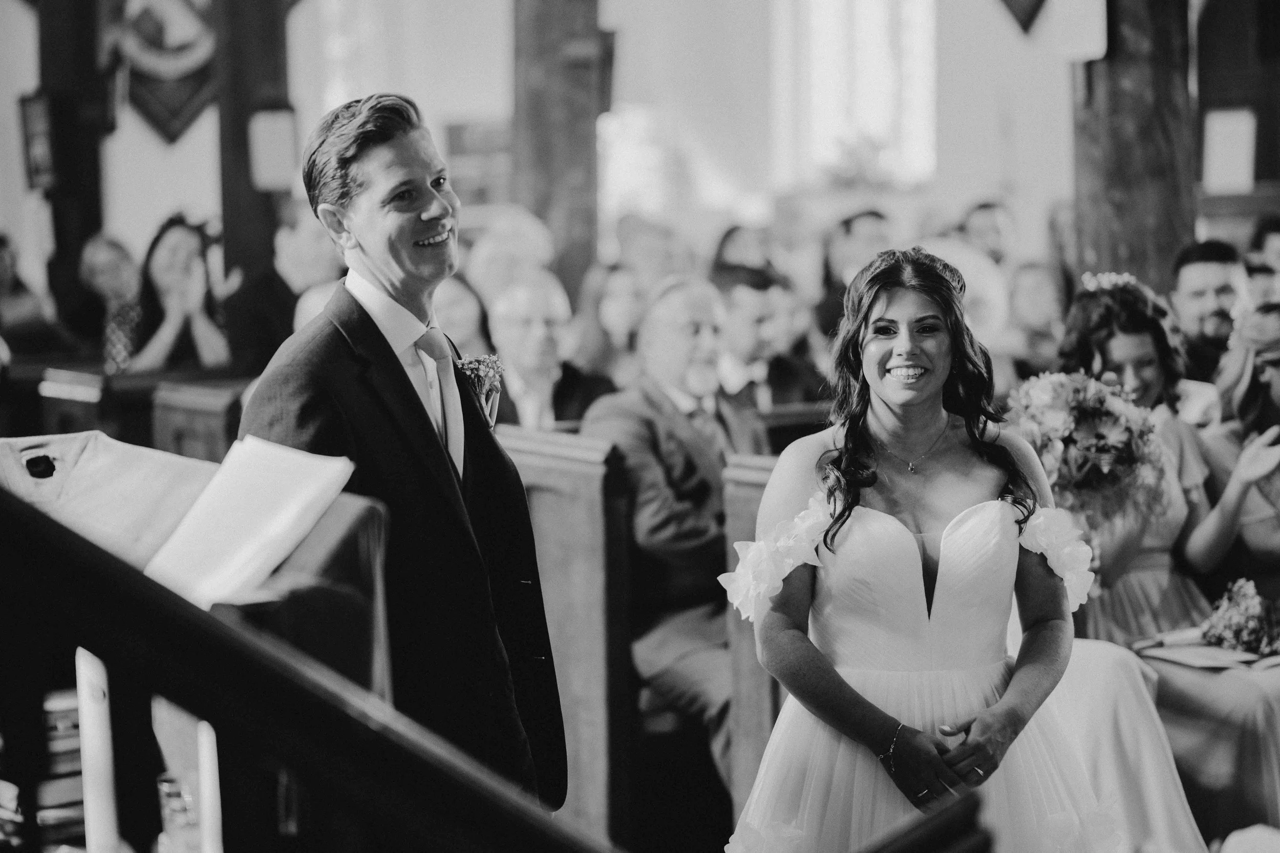 Black and white photo of a wedding ceremony inside a church with a smiling bride and groom standing at the altar, surrounded by guests.