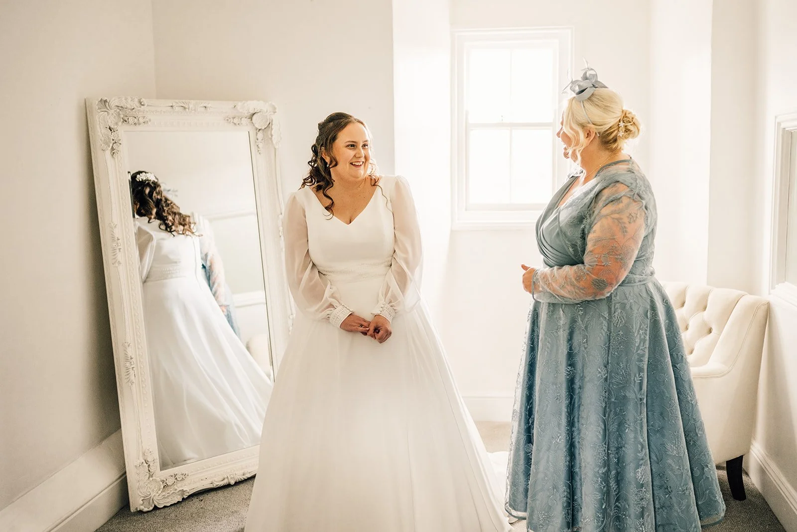 A bride in a white wedding gown smiling and talking with a woman in a blue floral dress, inside a bright room with a large mirror and a window.