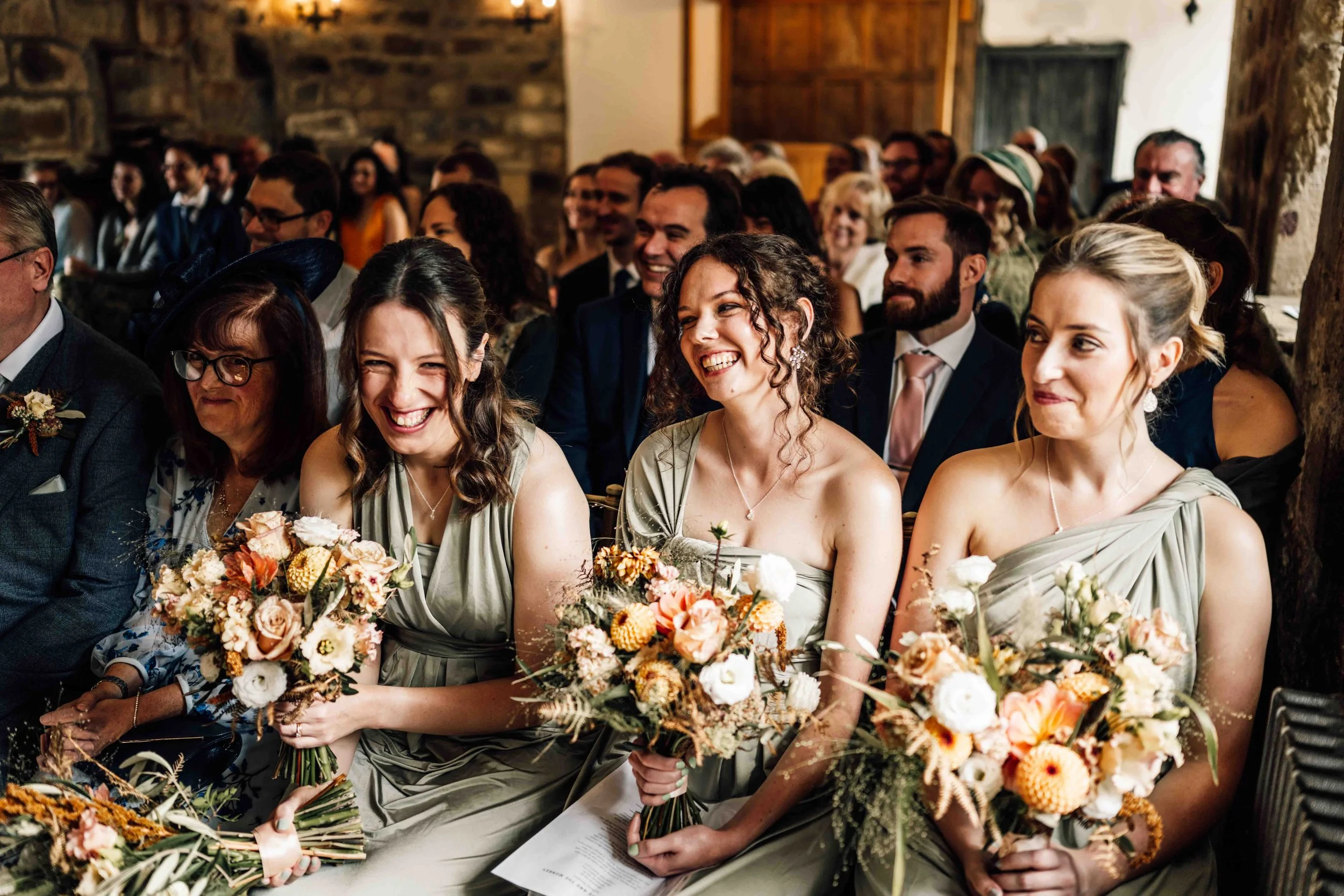 A group of women and men dressed in formal attire, sitting in a rustic wooden and stone venue, attending a wedding ceremony. Three women in light-colored dresses holding bouquets are in the front row, smiling and laughing.