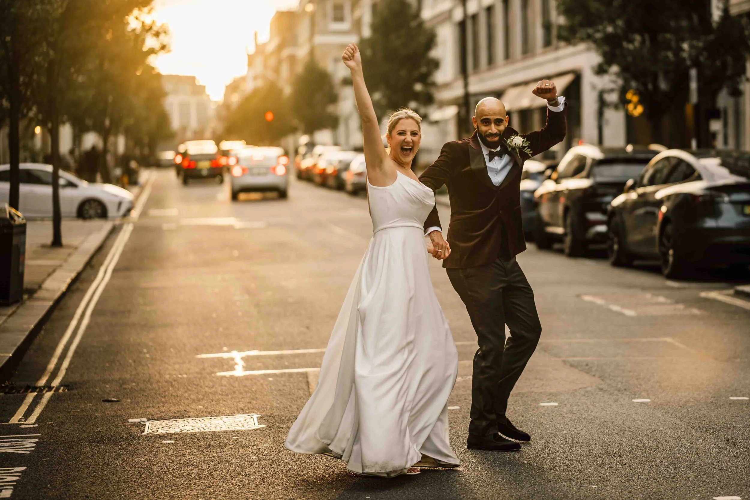 A smiling bride and groom celebrating on a city street at sunset, with the bride in a white wedding dress and the groom in a dark tuxedo, holding hands and raising their fists in celebration.