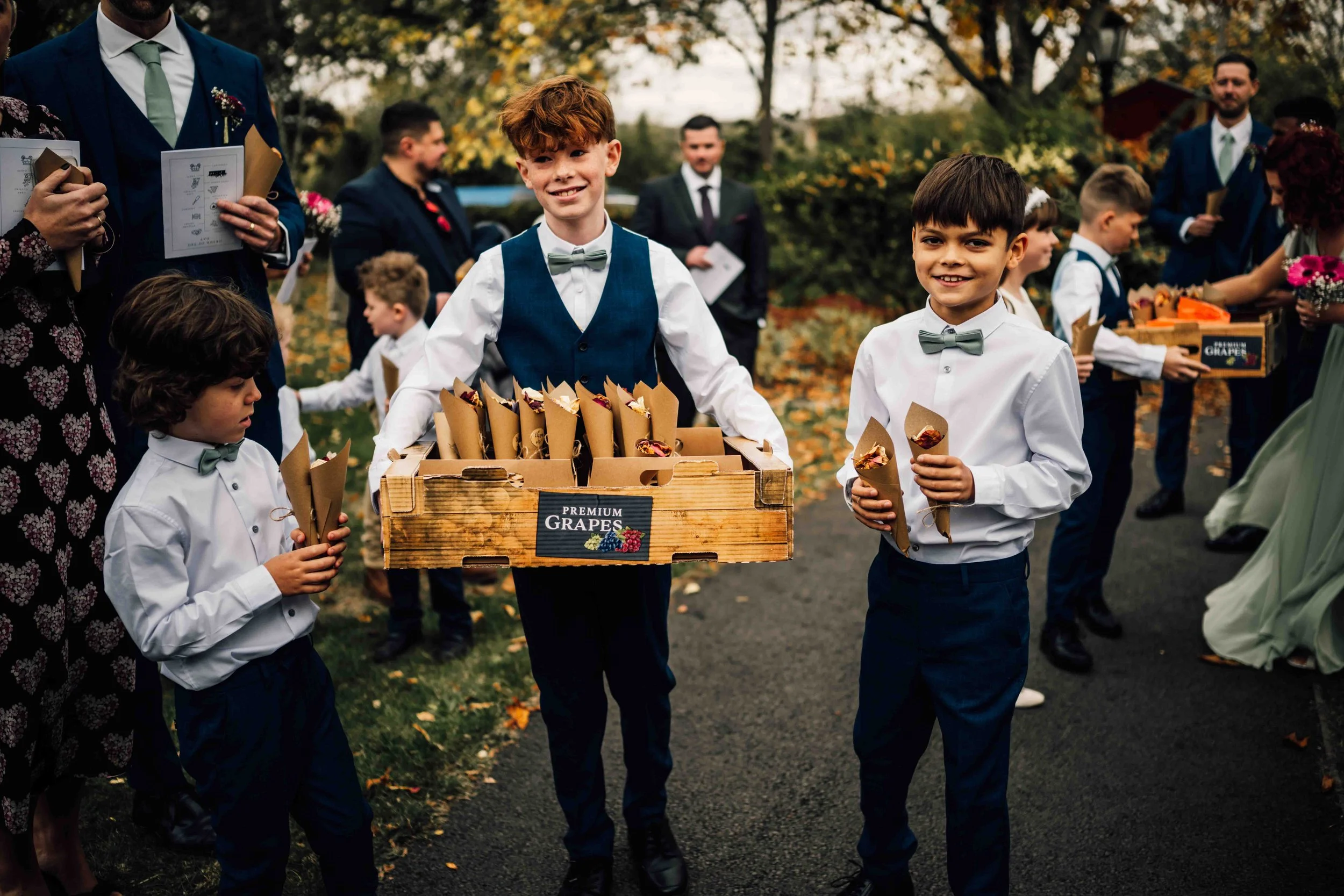 Young boys at a wedding holding cones of food, one boy in the center carrying a box labeled 'Premium Grapes.'