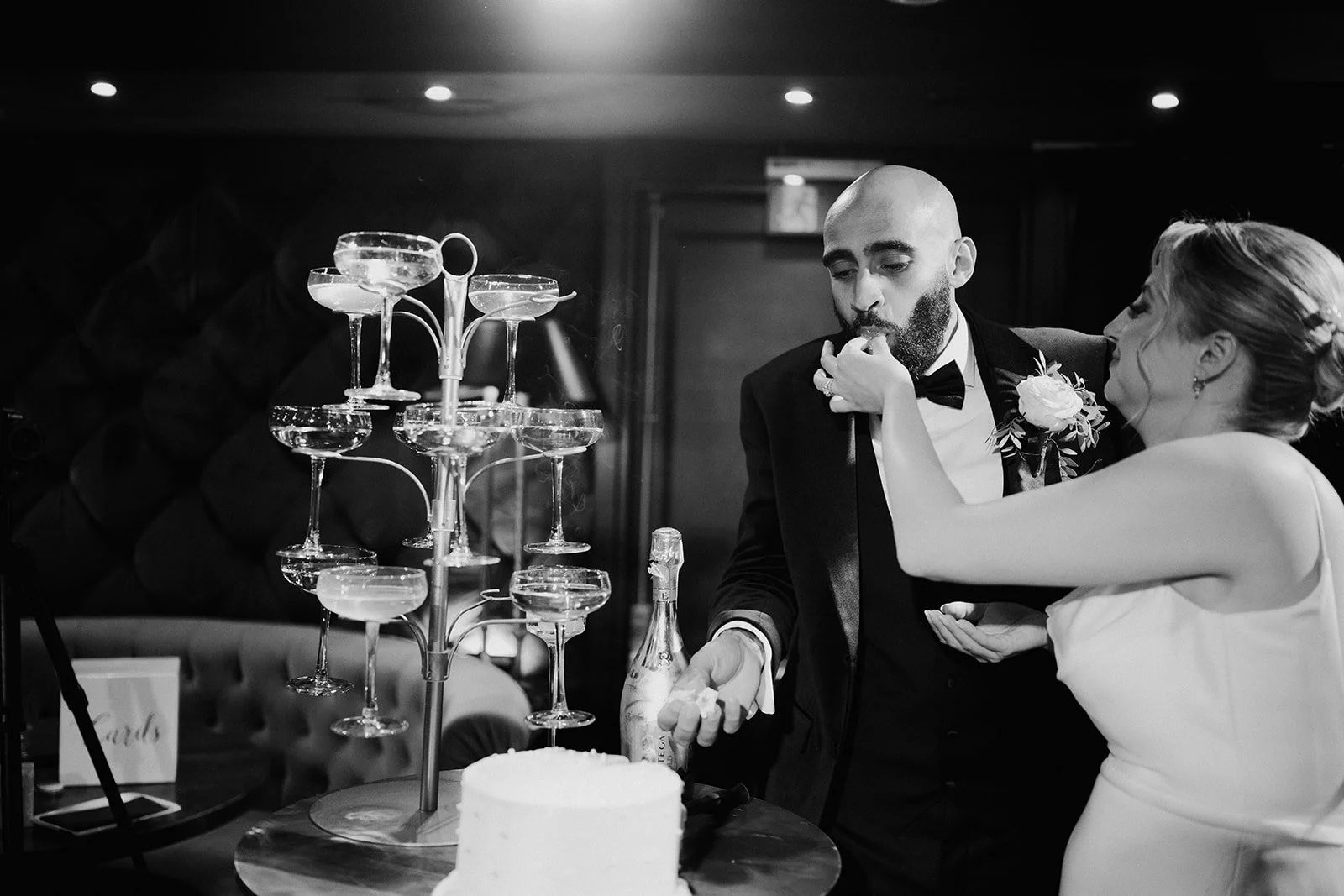 A couple at a wedding reception, with the woman feeding the man a piece of cake, surrounded by champagne glasses and a wedding cake on the table.