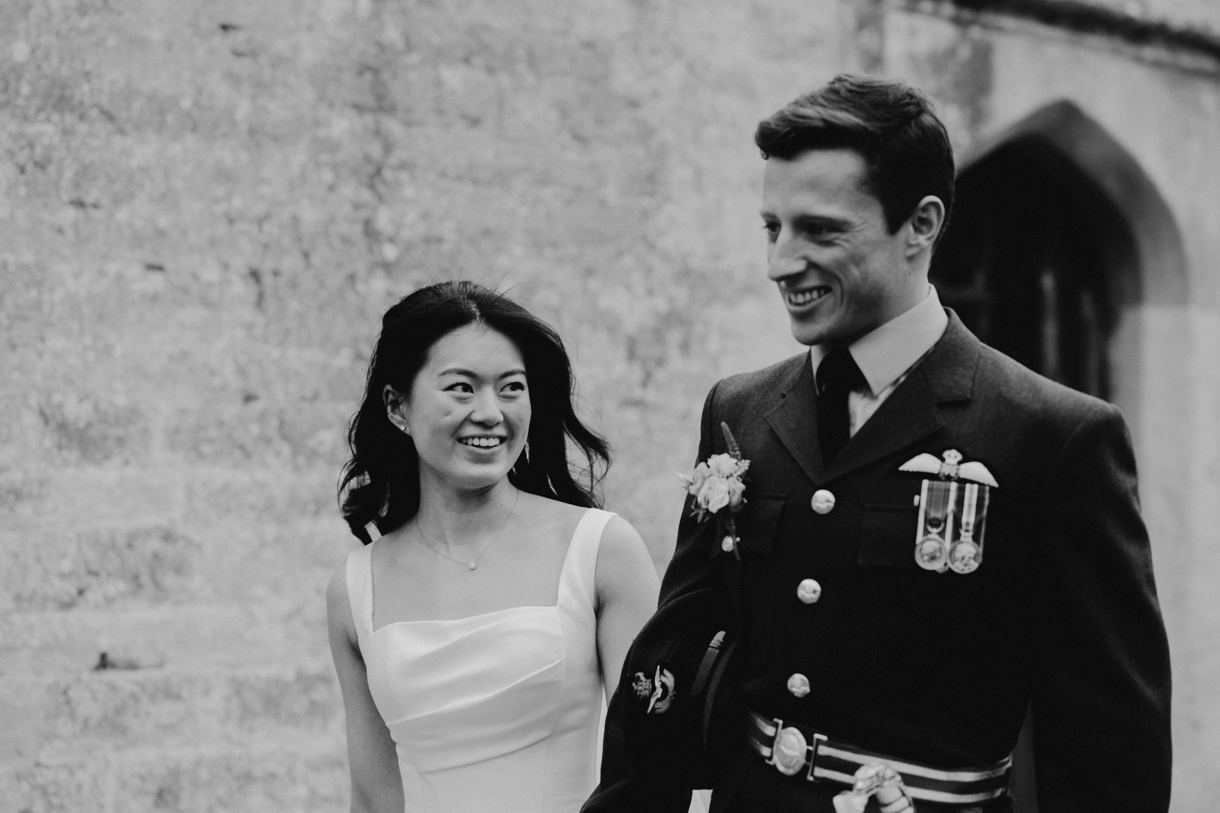 Black and white photo of a smiling woman in a white dress with an updo hairstyle and a man in a formal military uniform with medals and ribbons, standing outside near a stone building.