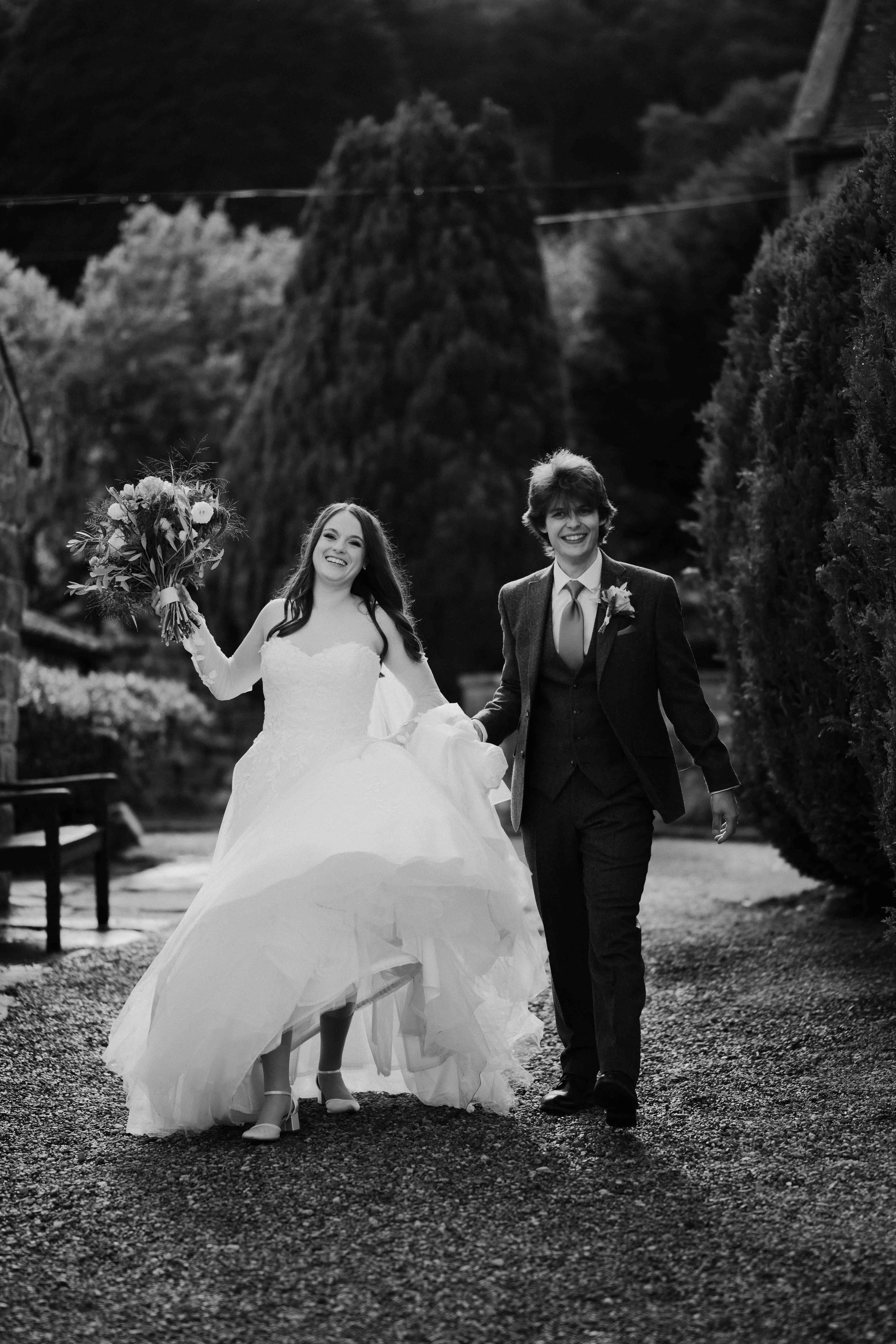 A black and white photo of a newlywed couple walking outdoors. The bride is holding a bouquet and lifting her dress, smiling. The groom is dressed in a suit with a boutonniere, also smiling. They are walking along a path with trees and bushes in the background.