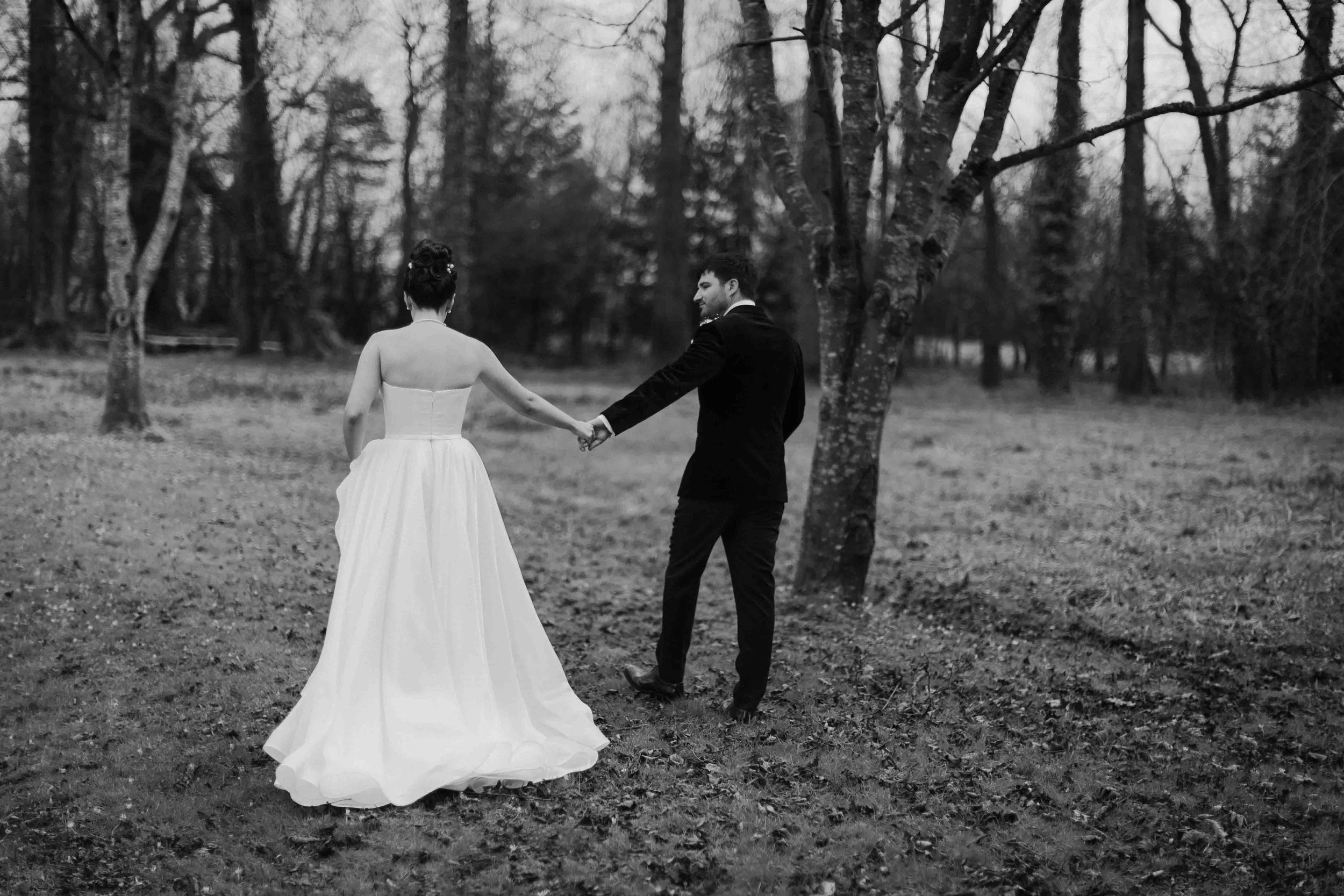 A black and white photo of a bride and groom holding hands and walking in a forested area. The bride is in a strapless wedding dress and the groom is in a dark suit. They are facing away from the camera, surrounded by leafless trees.