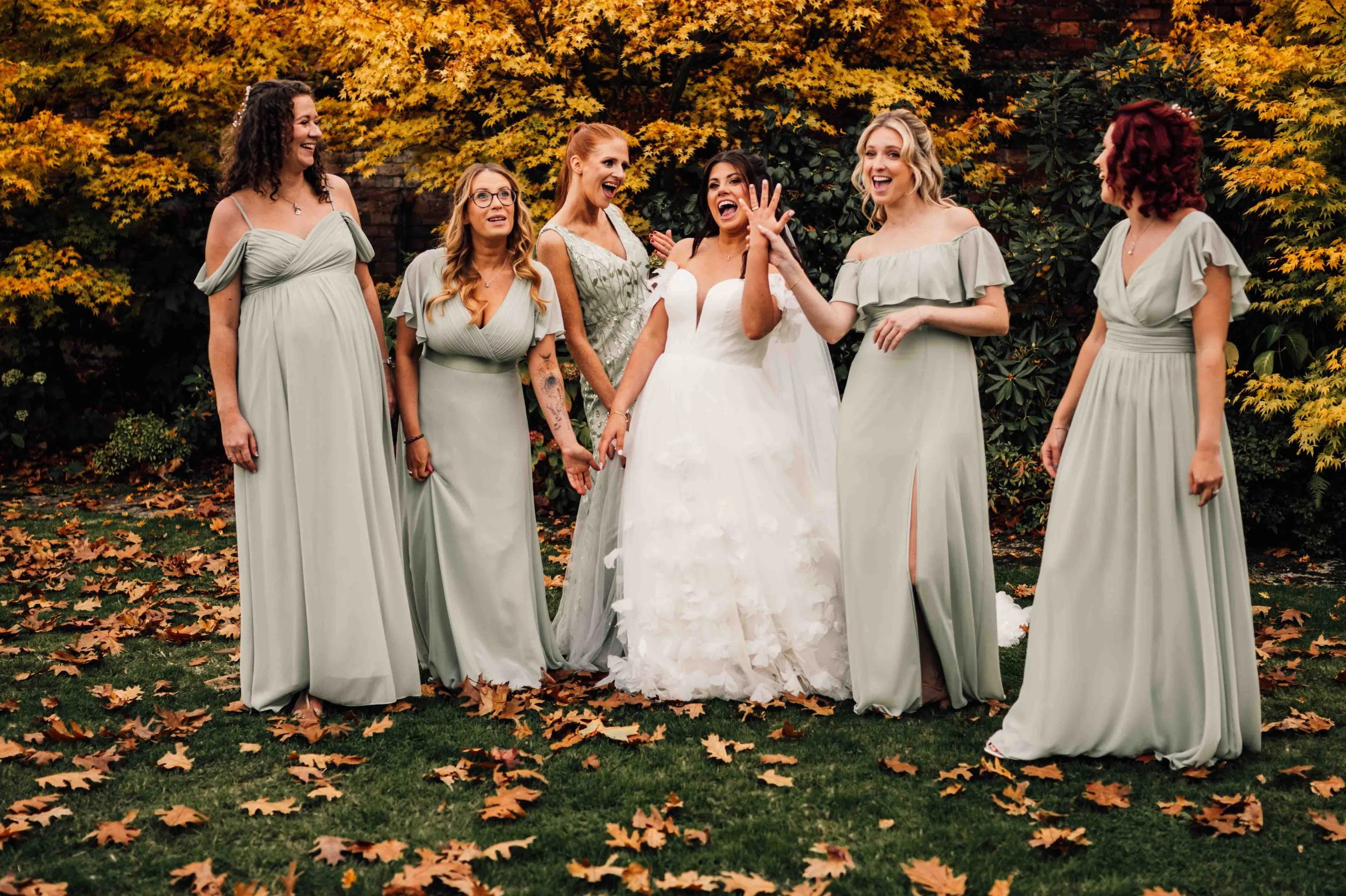 A group of seven women, including a bride in a white wedding gown and six bridesmaids in matching teal dresses, laughing and holding hands outdoors with autumn leaves on the grass and yellowish-orange foliage in the background.