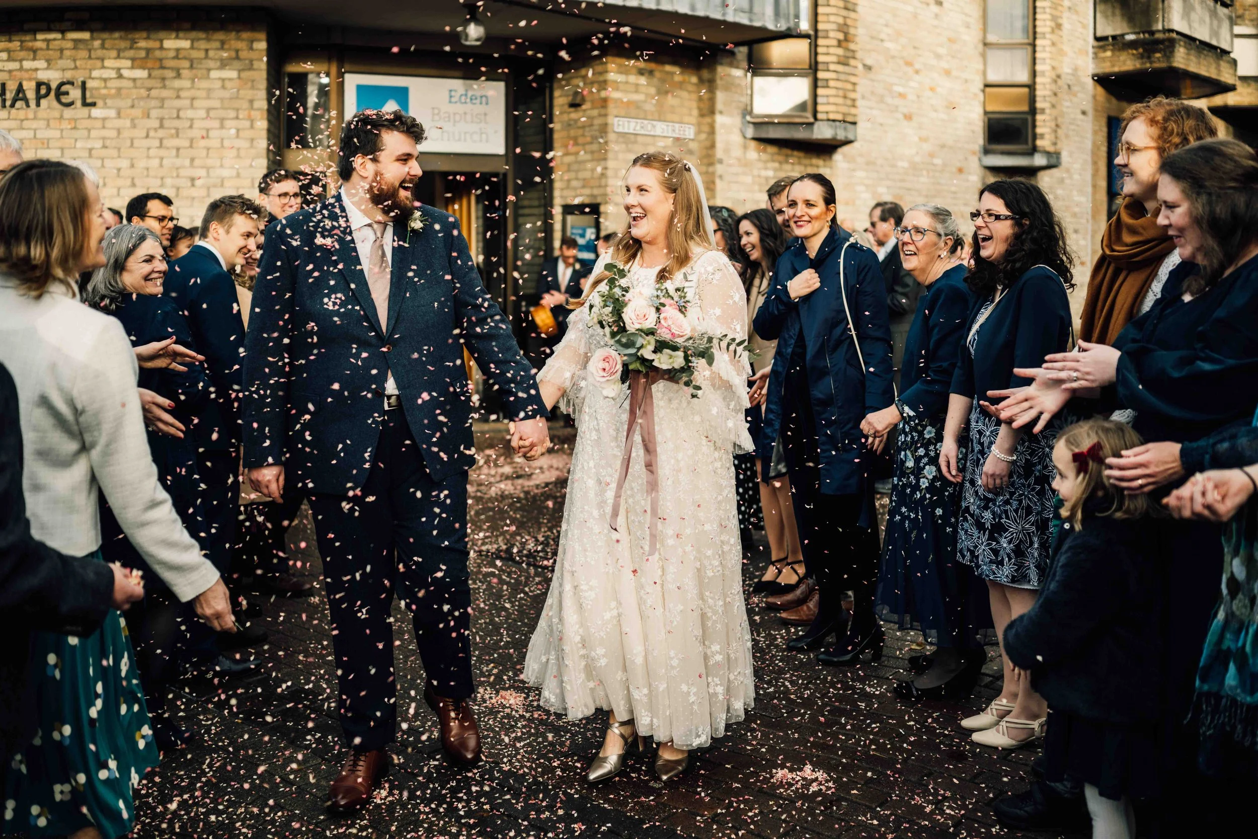 Bride and groom holding hands and smiling as they walk through a shower of pink confetti, surrounded by friends and family outside a church.
