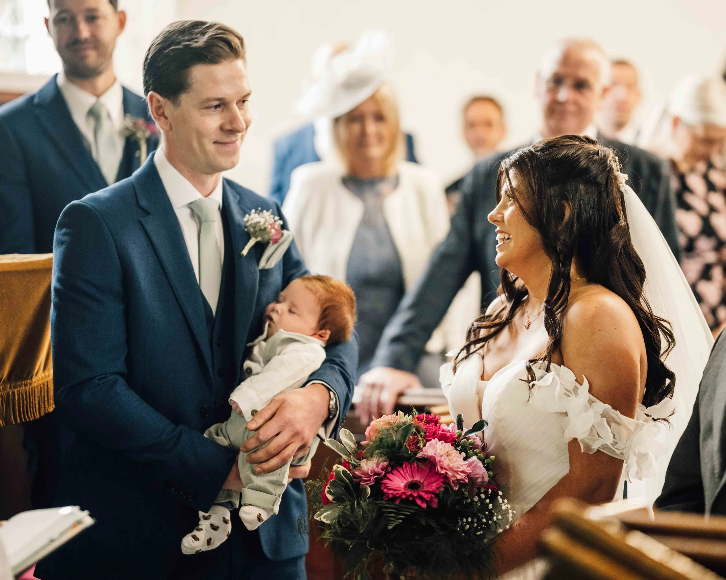 A groom holding a sleeping baby on his arm exchanging vows with a bride holding a bouquet during a wedding ceremony. Several guests are in the background.