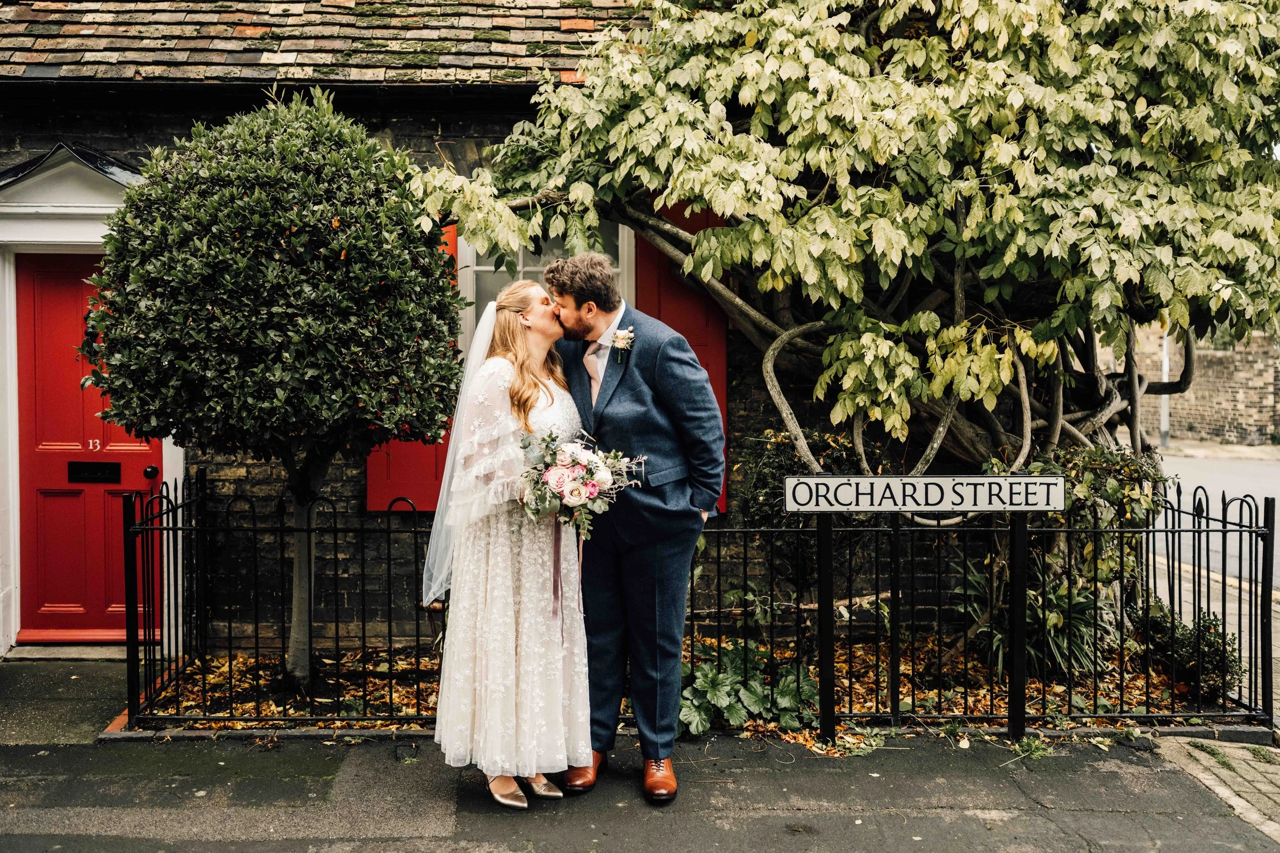A bride and groom kiss on a sidewalk near a red door and a Garden Street sign, with trees and greenery in the background.