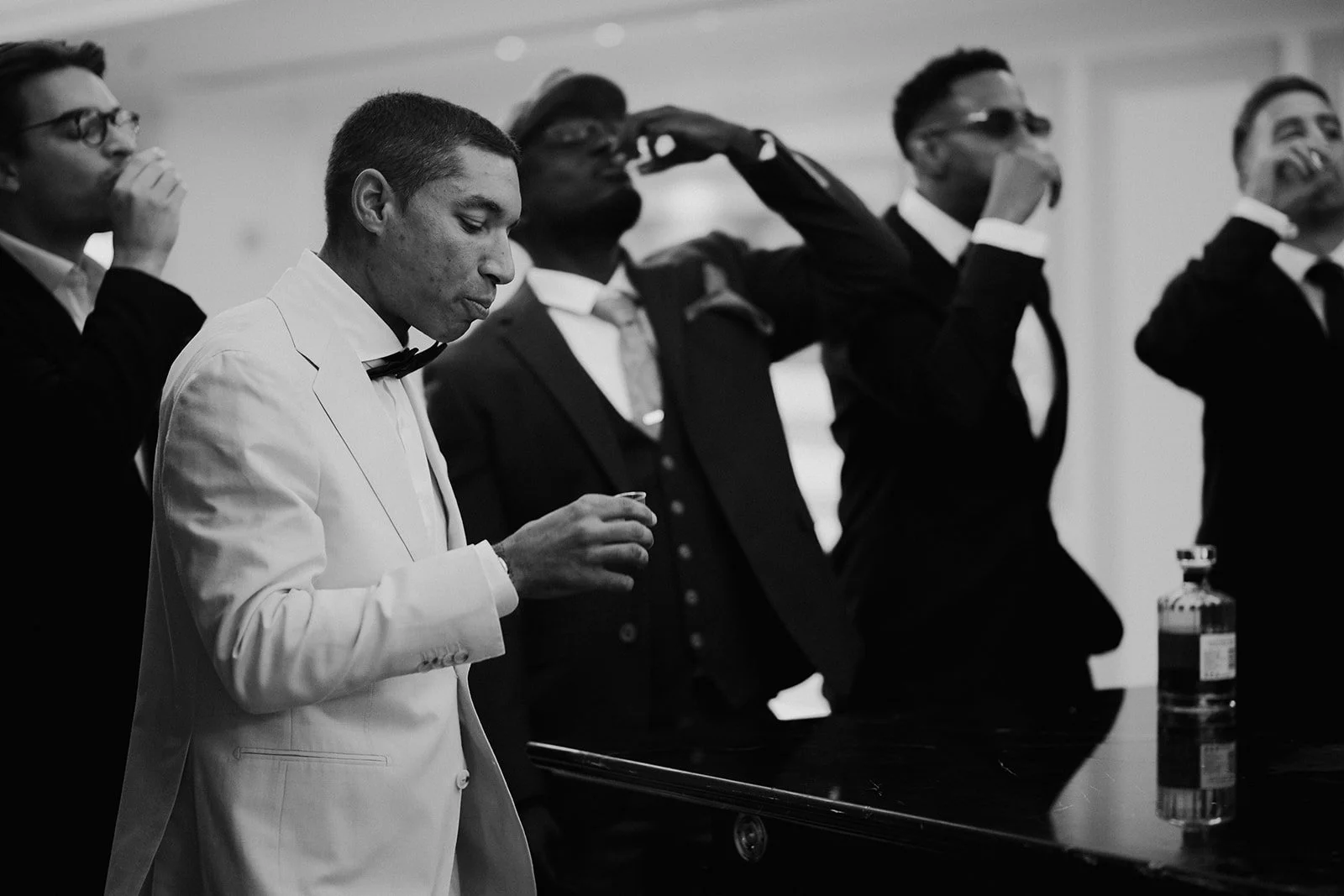 A black and white photo of five men dressed in formal tuxedos, with one in a white tuxedo jacket, standing at a bar or counter, holding glasses, with one man drinking from a glass and one with sunglasses, captured in a social or celebratory setting.