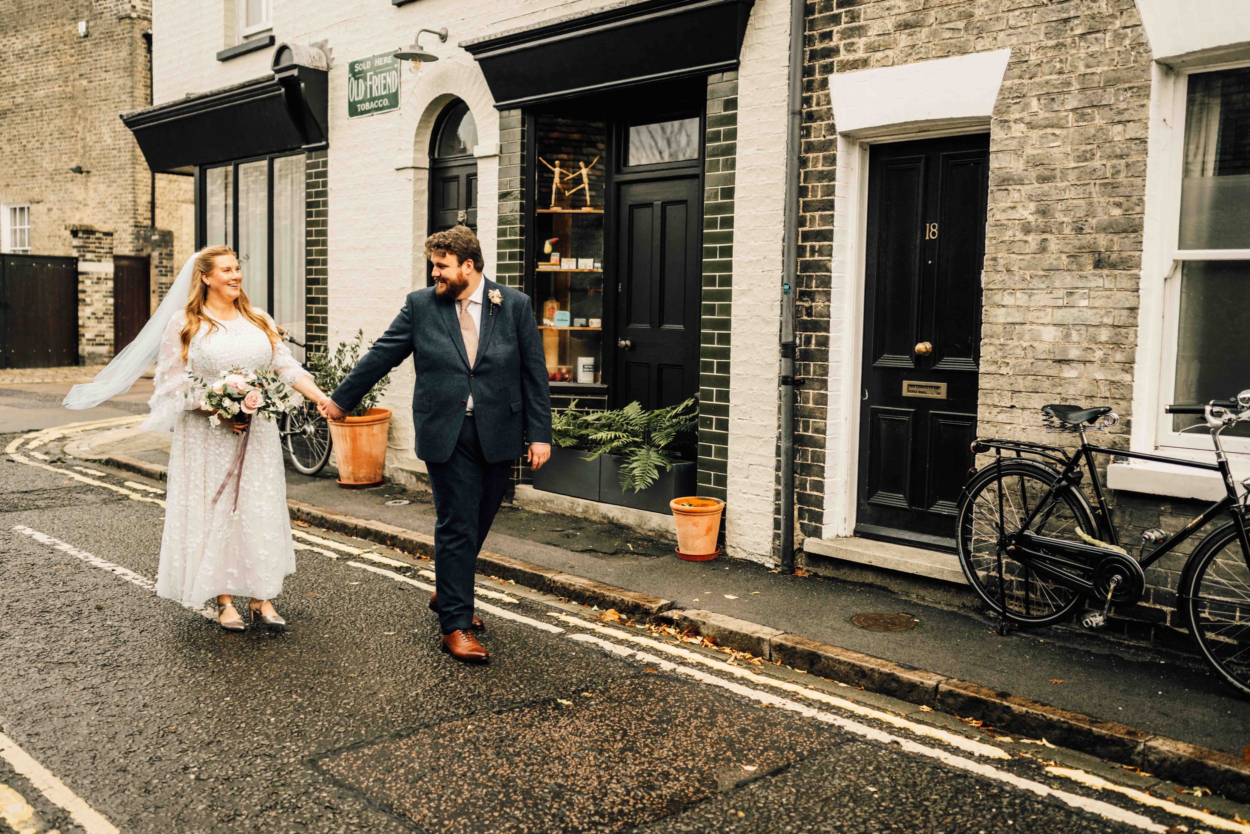 A couple in wedding attire walking hand in hand on a city street, with the bride in a white dress and the groom in a suit, smiling at each other.
