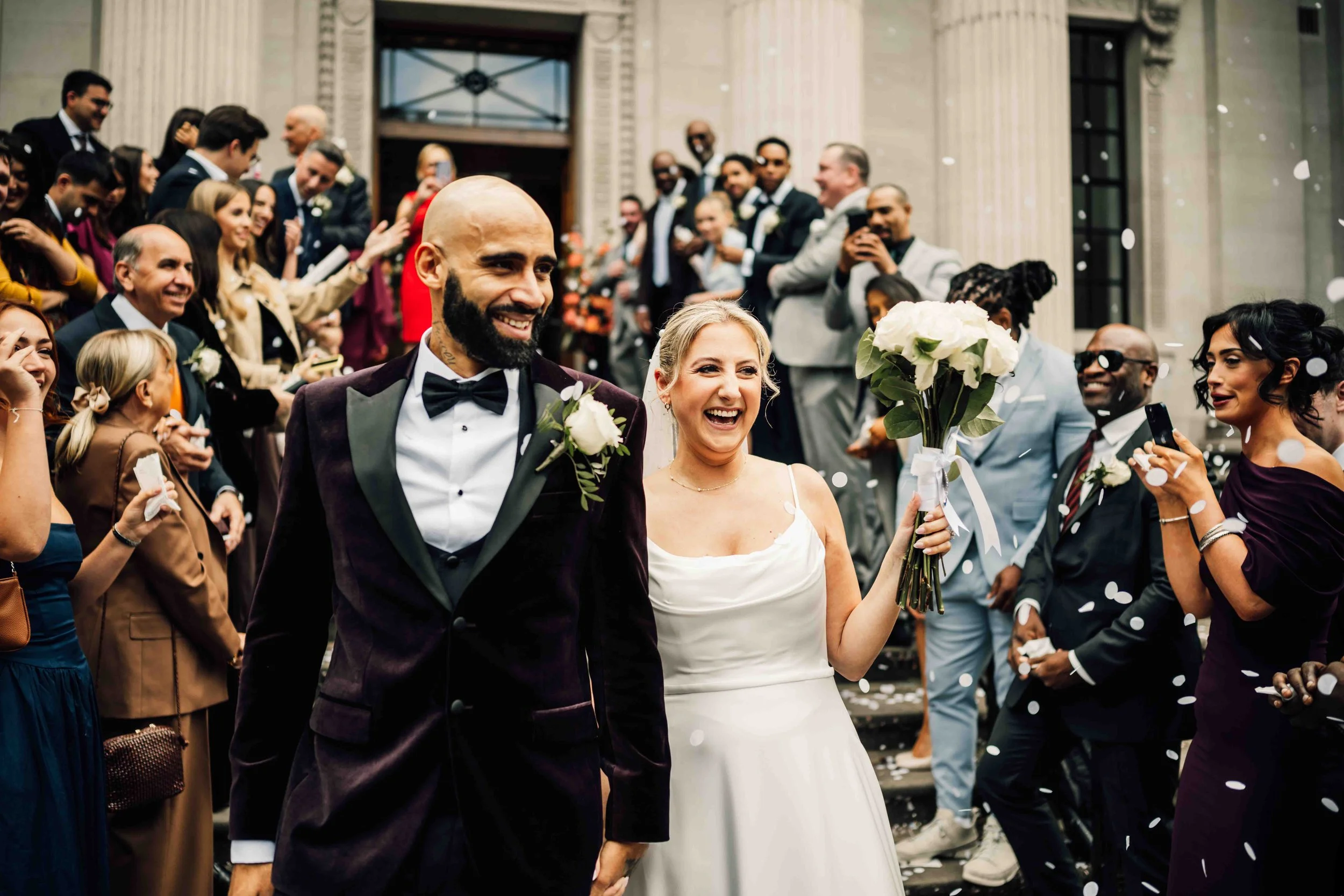 A newlywed couple is smiling as they leave a building, holding flowers, while surrounded by friends and family celebrating with confetti.
