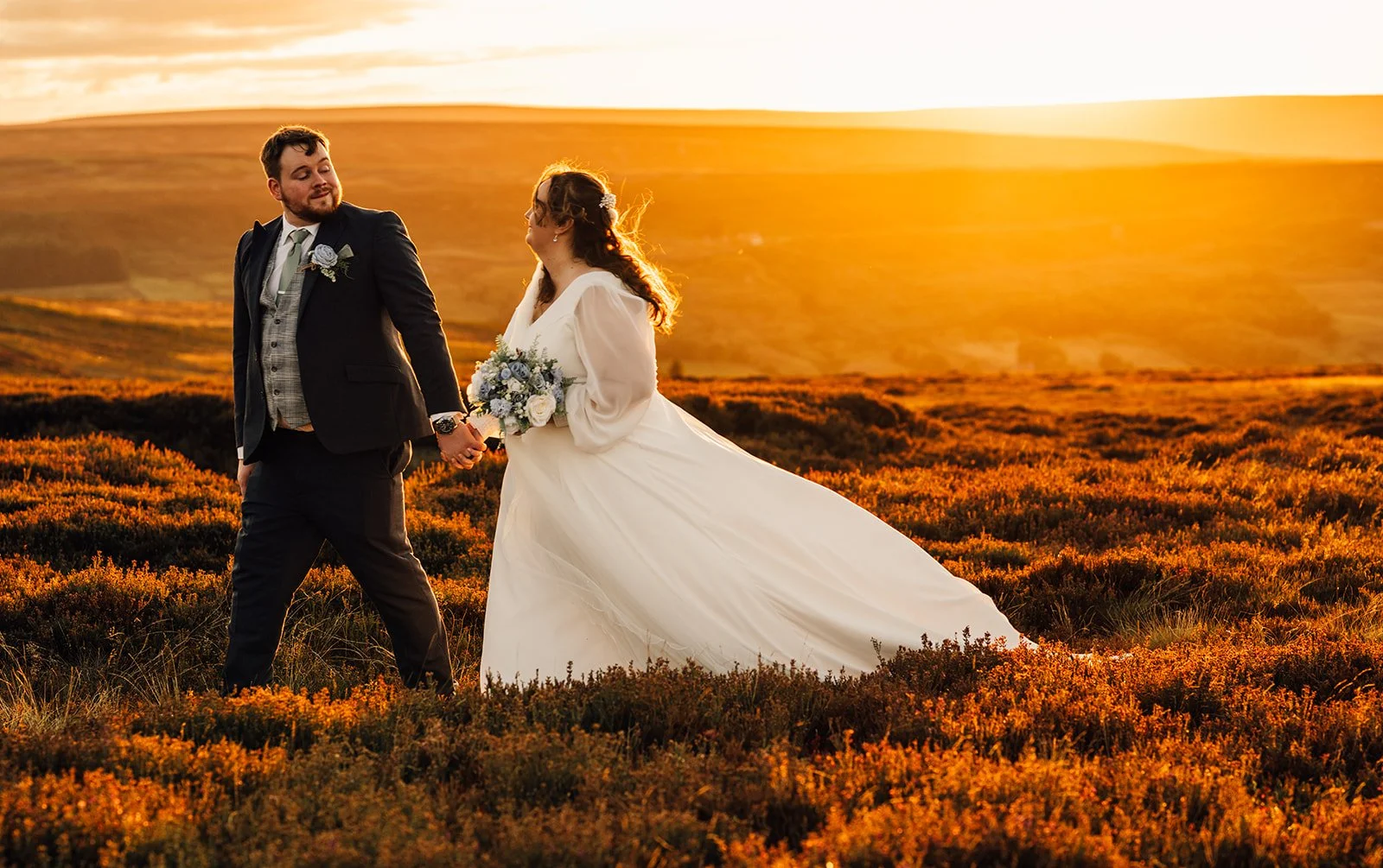 A bride and groom holding hands in a sunset landscape, with the bride wearing a white gown and holding a bouquet, and the groom in a dark suit with a vest and tie, standing amid orange and green vegetation.