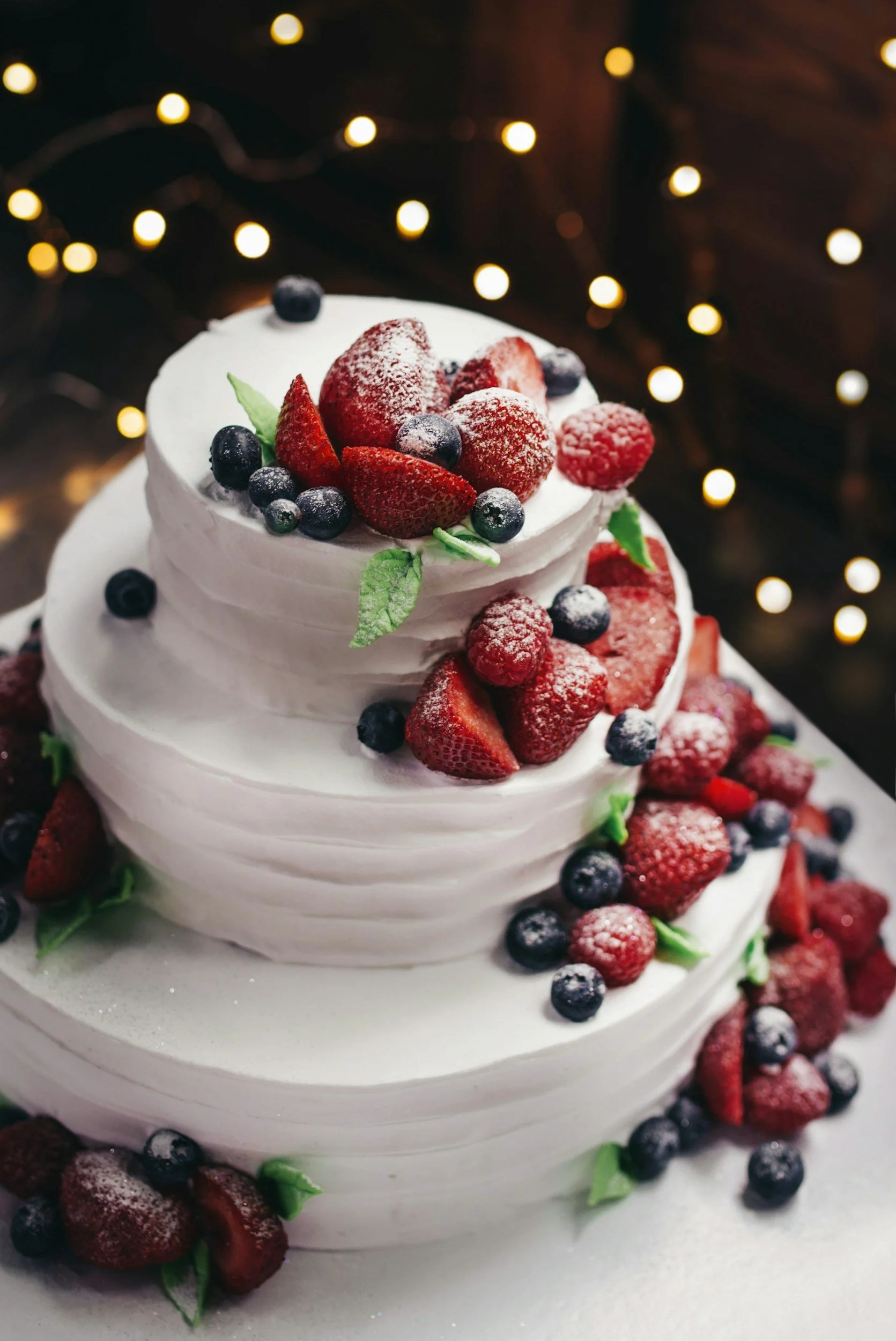 A three-tiered white frosted cake decorated with fresh strawberries, blueberries, raspberries, and green leaves, with a blurred string of warm yellow fairy lights in the background.