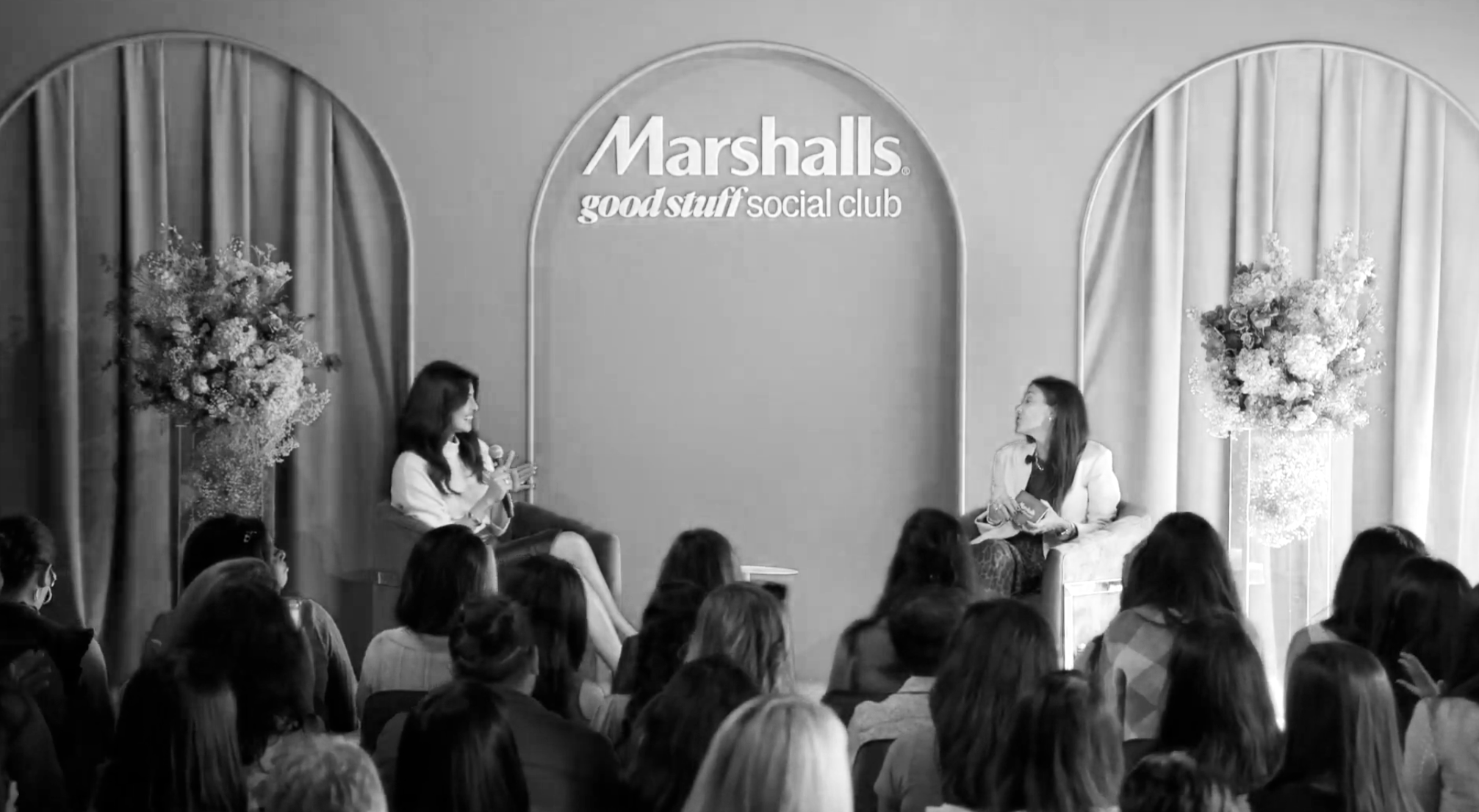 Two women are seated on stage in front of an audience at the Marshalls social club, with a sign on the wall behind them reading "Marshalls good stuff social club." There are large floral arrangements on either side of the stage.