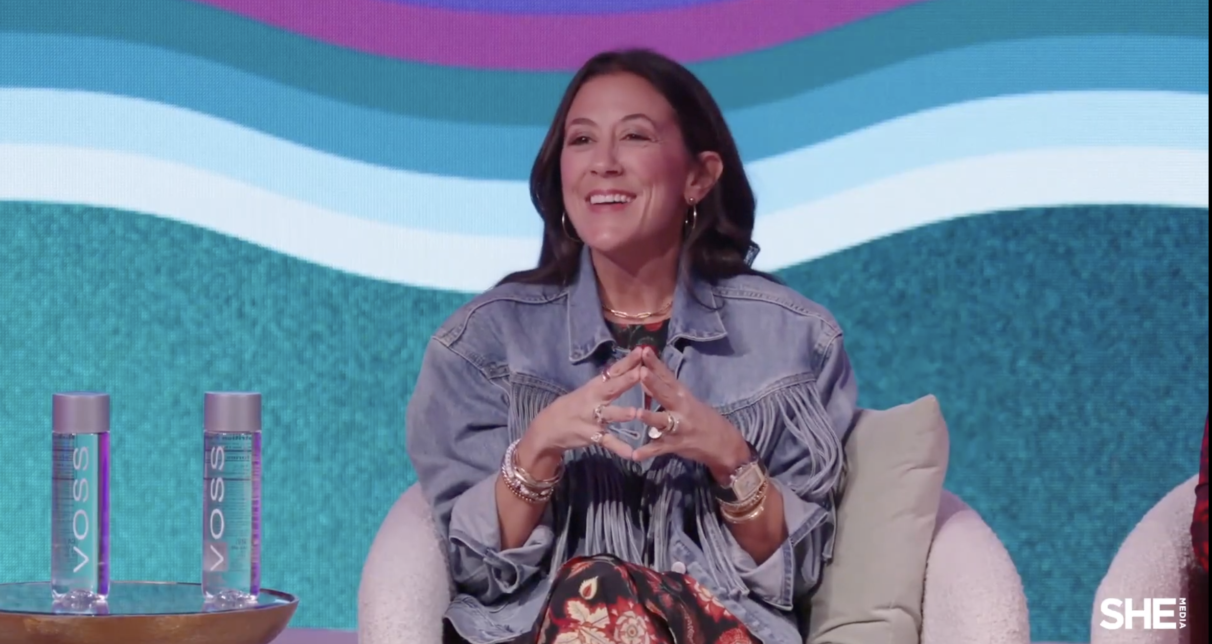 A woman with dark hair, wearing a denim jacket and colorful patterned pants, smiling and sitting on a beige armchair with her hands clasped together. There are two bottles of VOSS water on a tray to her left, with a colorful wavy background behind her.
