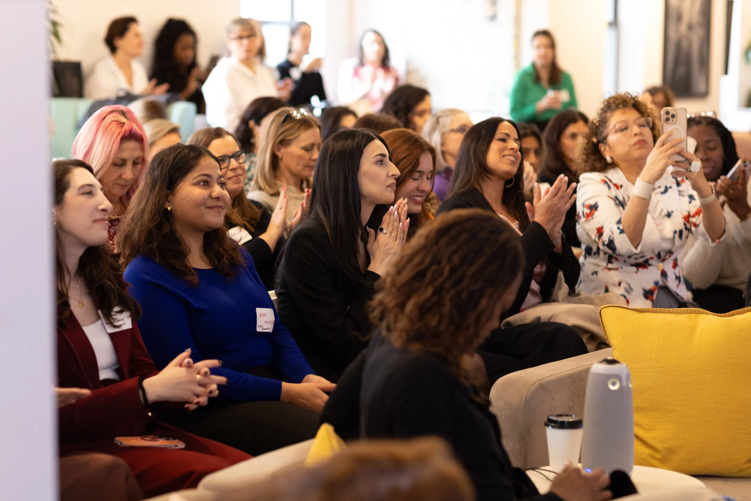 Women seated in an audience at a conference or seminar, some clapping and taking photos.