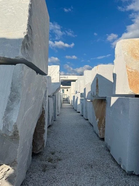Massive white marble blocks line a narrow gravel pathway at a stone yard beneath a bright blue, partly cloudy sky.